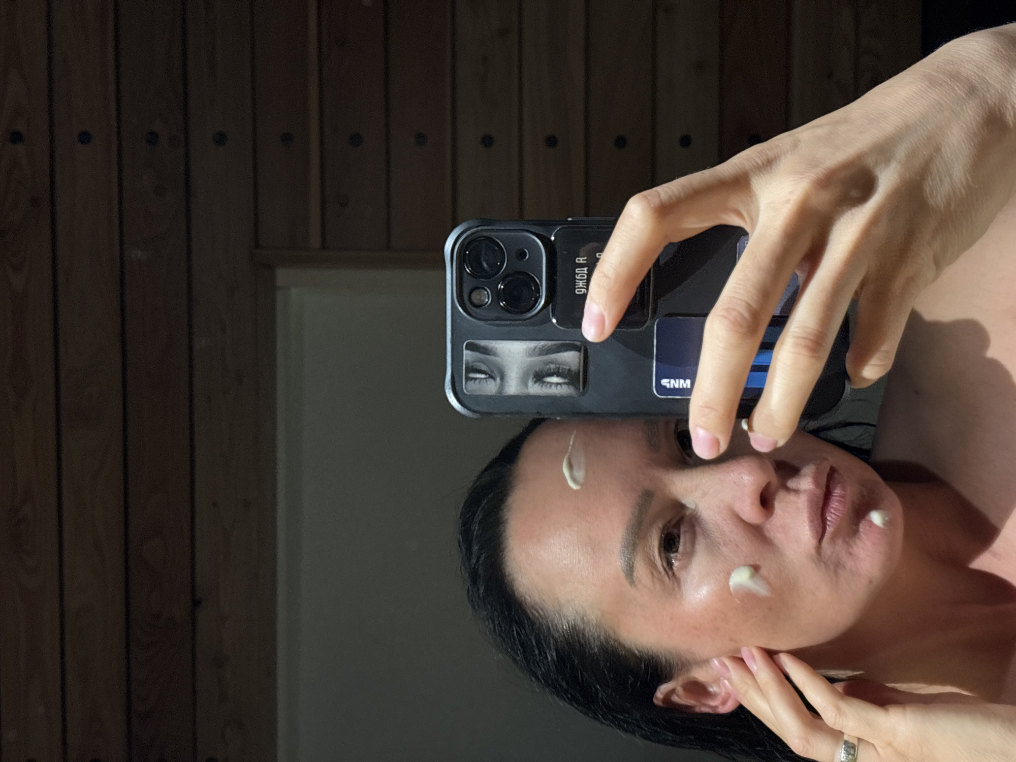 A person is taking a selfie in front of a wooden paneled wall, likely a mirror reflecting the panels. The person is applying cream to their face, with visible dabs of white cream on their forehead, cheek, and around their mouth. They are holding a smartphone with a distinctive case, featuring a graphic of eyes and other design elements, indicating it may be a personalized or branded case. The image appears to be taken indoors. The location cannot be determined from the given image.