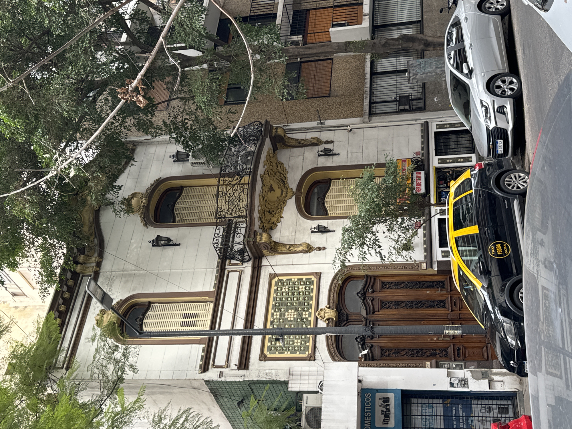 The image shows a street scene in Buenos Aires, Argentina, with a focus on a building featuring ornate, classical architectural details. The building is cream-colored with detailed woodwork around the windows and doors. A wrought-iron balcony is visible on the upper level. A taxi, partially visible, is parked in front of the building. A silver car is parked behind it. Street-level businesses, including a store labeled "DOMESTICOS" and a hardware store with a "FERRETERIA" sign, occupy the lower part of the building. Trees frame the building and line the street.