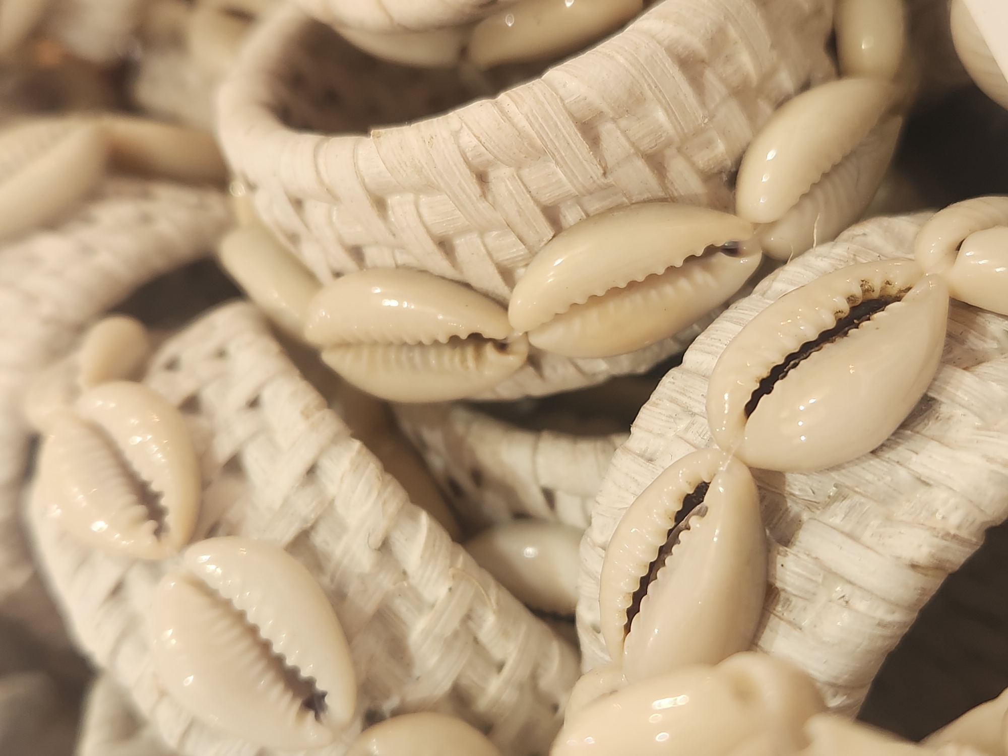This media file showcases a close-up, shallow-depth-of-field view of several decorative woven items adorned with cowrie shells. The objects appear to be circular or cylindrical in shape, possibly napkin rings or small decorative baskets, crafted from a light-colored, woven material, likely straw or raffia. The shells, natural and creamy white with dark openings, are attached along the exterior of these woven objects, creating a textured and eye-catching design. The lighting suggests an indoor setting, possibly a craft market or a home decorated with coastal-themed items. There is no discernible text within the frame. The image focuses on the intricate details of the weaving and the organic shapes of the shells, emphasizing their decorative qualities. The scene is still and offers no indication of specific time of day, weather, or emotional context beyond a sense of artisanal craftsmanship. The location context provided is Maitland, United States of America, which could imply the origin or sale of such decorative items.