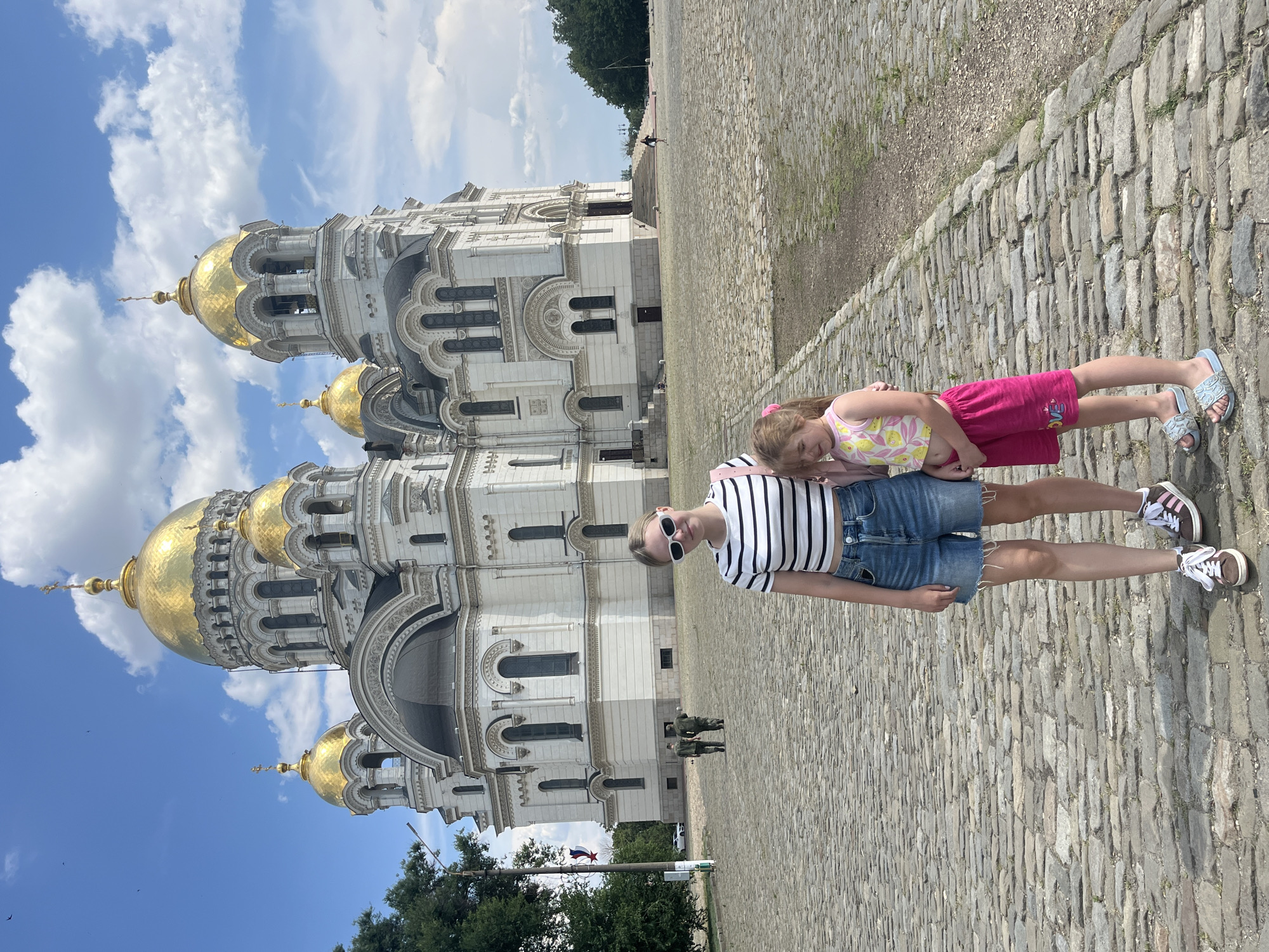 Two young females, one older and one younger, stand on a cobblestone plaza in front of a large white cathedral with golden domes. The older female, on the left, wears a striped t-shirt, denim shorts, white sunglasses, and brown and white sneakers. The younger female, on the right, wears a pink floral tank top, pink shorts, and blue slides. The cathedral, identified as the Ascension Cathedral in Novocherkassk, Russia, features multiple domes and ornate architectural details. In the background, two individuals in military uniforms walk away from the camera, and other people can be seen in the distance near the entrance of the cathedral. Trees line the left side of the image, and a flagpole with a Russian flag is visible. The sky is blue with scattered clouds.