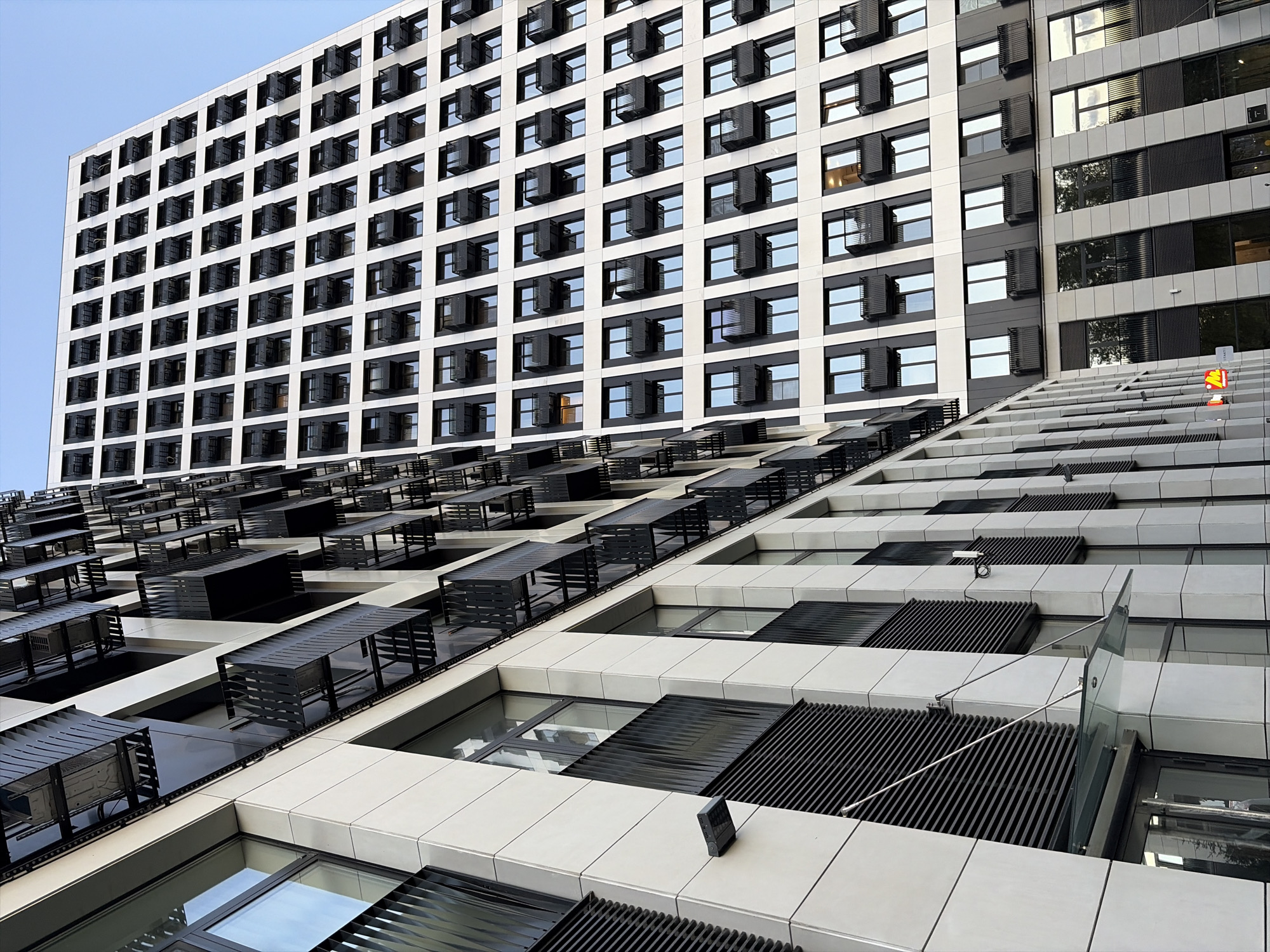 The photograph captures a low-angle perspective of two modern apartment buildings in Moscow, Russia. The building in the foreground features a light-colored facade with numerous windows and black balconies. To the right, a taller building with a similar aesthetic rises, its facade predominantly white with a grid-like pattern of dark balconies and windows. The sky above is a clear, light blue. The architecture suggests a contemporary urban development.