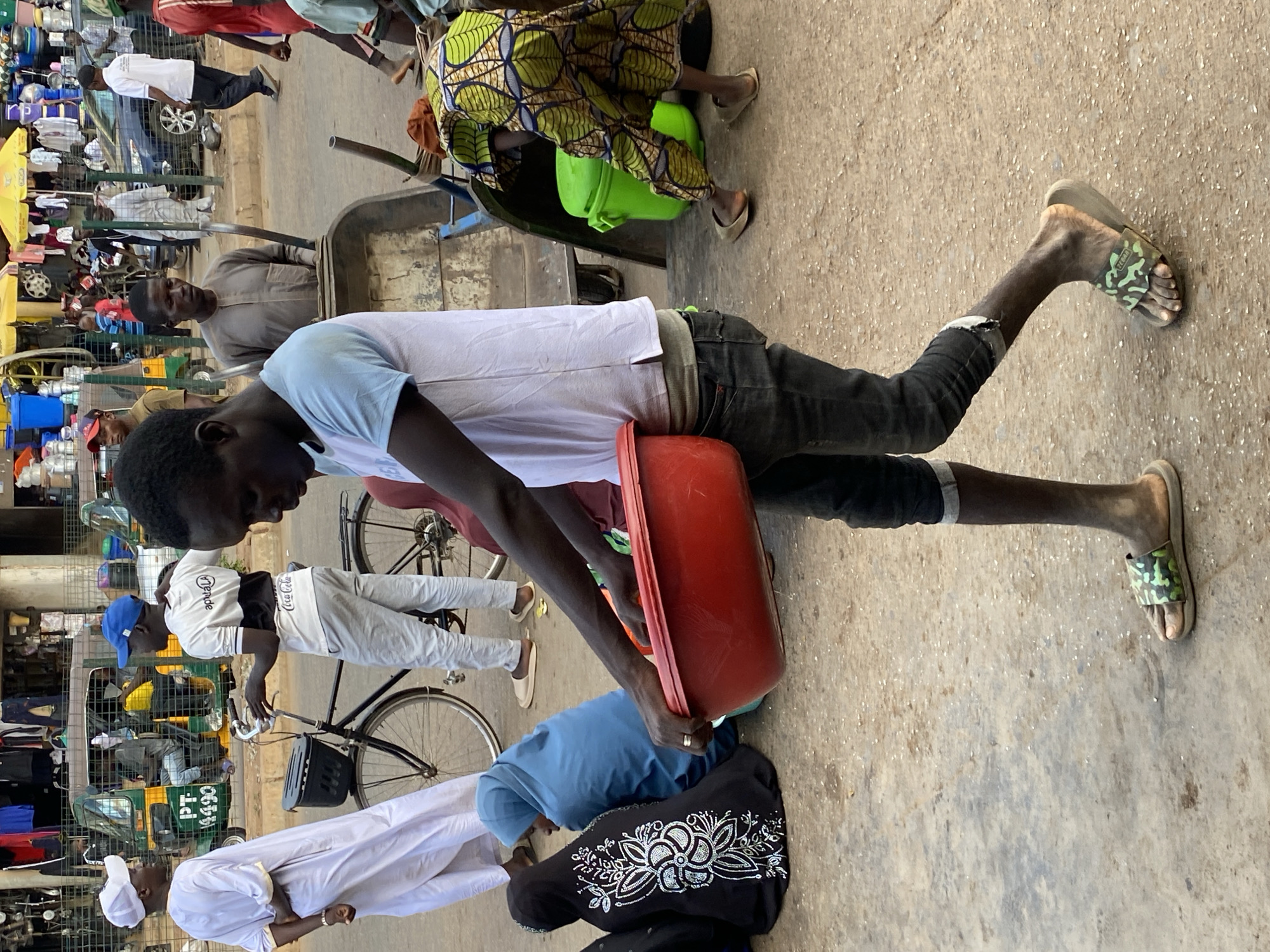 In Bukuru, Nigeria, the scene depicts a bustling street. A person wearing a white t-shirt and dark jeans carries a red bucket. Nearby, individuals in various attire are present. One person is wearing a green patterned cloth, while another is bending over in a blue top. There are also people in white robes and head coverings, along with others riding bicycles. A cart filled with items is visible, and a man wearing a brown robe stands near the cart. Several shops with various merchandise and a yellow tuk-tuk are also present.