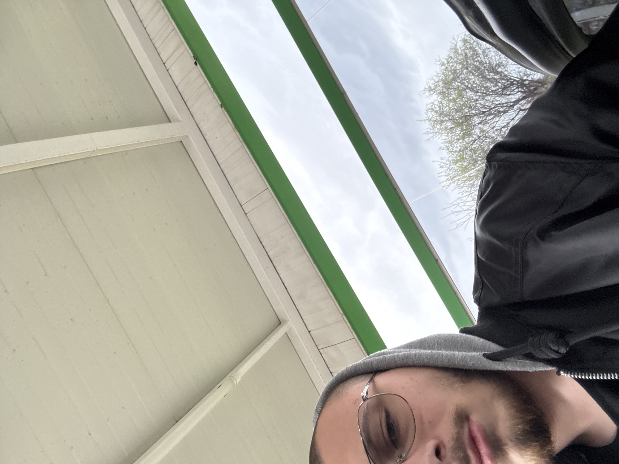 The image is a low-angle, upward-looking selfie of a person wearing glasses and a grey beanie, with part of their face and beard visible. They are also wearing a black jacket. The background consists of a white corrugated metal structure with green support beams, above which is a cloudy sky and the bare branches of a tree. The location is a covered area, potentially a gas station or similar structure. The photo was taken outdoors in Stara Zagora, Bulgaria.