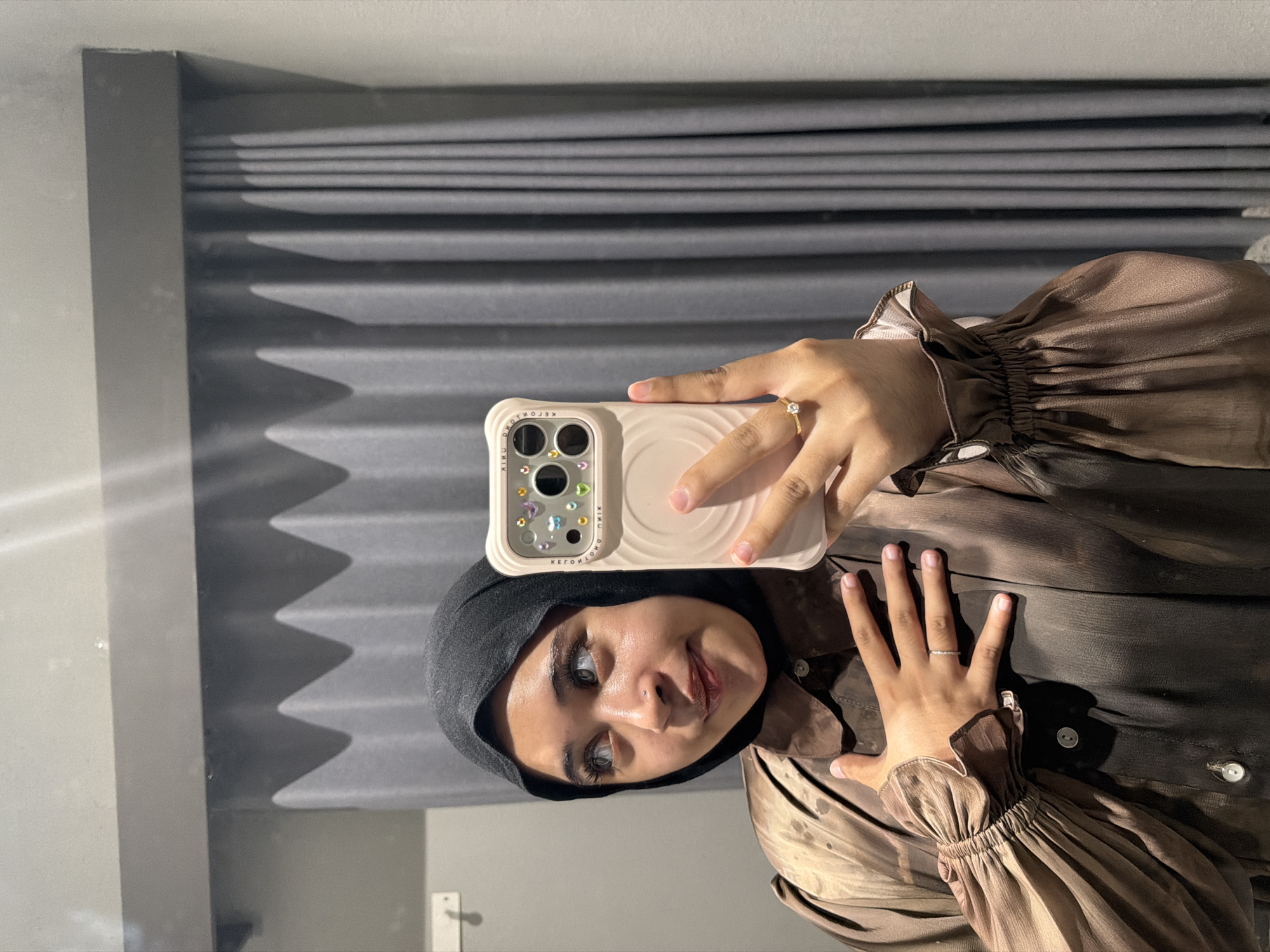 In a fitting room, a young woman wearing a black hijab and a brown silk blouse with puffed sleeves is taking a mirror selfie. She is holding a light pink phone decorated with colorful rhinestones in her right hand and has her left hand on her chest. A diamond ring is visible on her left ring finger. The background consists of grey pleated curtains and a gray wall with a light fixture. The overall impression is that of a casual moment captured in a private space.