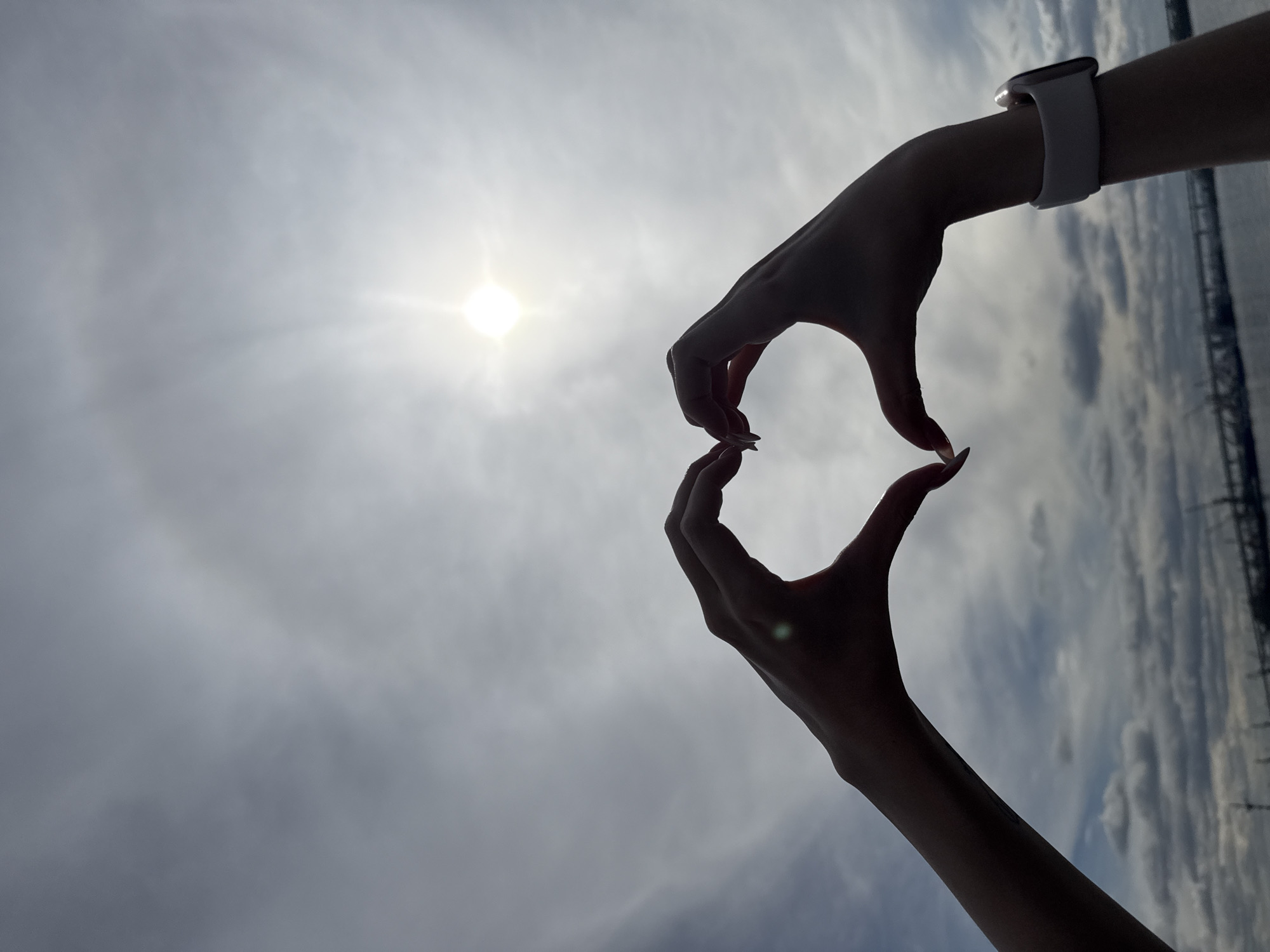The image displays two silhouetted hands forming a heart shape against a bright, cloudy sky. The sun, a brilliant white orb with radiating light, is prominently featured in the upper left quadrant, creating a halo effect. On the left wrist, a white smartwatch with a light-colored band is clearly visible. A small, sparkling object, possibly a ring or engagement ring, is nestled between the fingertips at the base of the heart shape, catching the sun's glare. The fingers are long, and the nails appear manicured with a light polish.
The background primarily consists of diffuse clouds and a bright sky, indicative of daytime, likely mid-day or early afternoon. To the far right, a faint, dark silhouette of a structure, possibly a bridge or scaffolding, is visible against the lighter horizon. The gesture of forming a heart with hands typically conveys love, affection, or gratitude. This scene is set in Saint Petersburg, Russia, with the faint structure hinting at the city's architectural landscape, perhaps one of its many bridges. There is no visible text in the image.