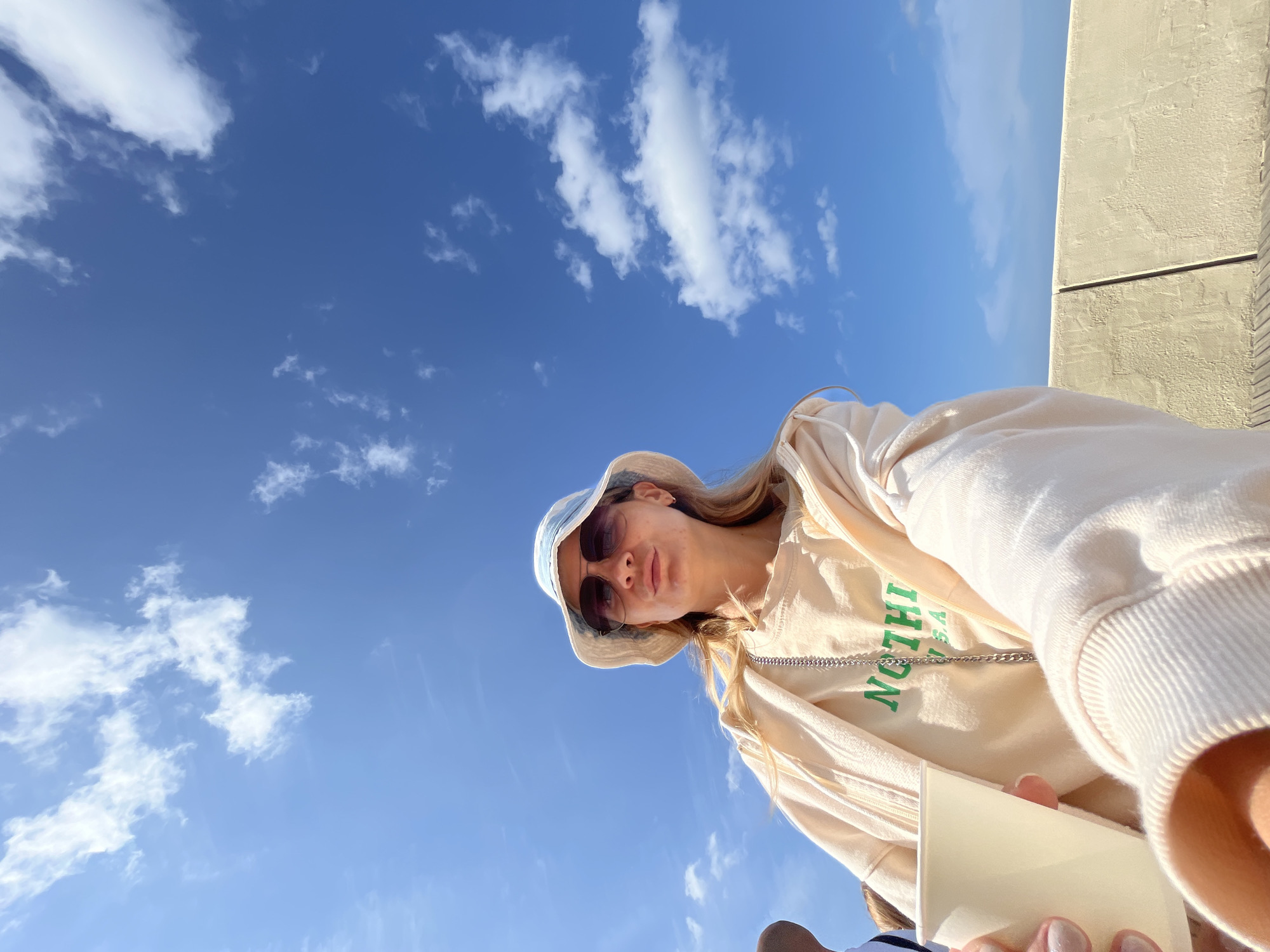 A woman wearing a bucket hat and sunglasses takes a selfie outdoors on a bright, sunny day with a blue sky and scattered clouds. She is holding a white cup in her left hand and is wearing a light-colored sweatshirt with green text that reads "NOTHING USA". She appears to be seated on a bench. The background consists of a textured wall and a wooden surface, suggesting an outdoor seating area. The image does not provide specific visual cues to confirm the location as Svetlogorsk, Russia.