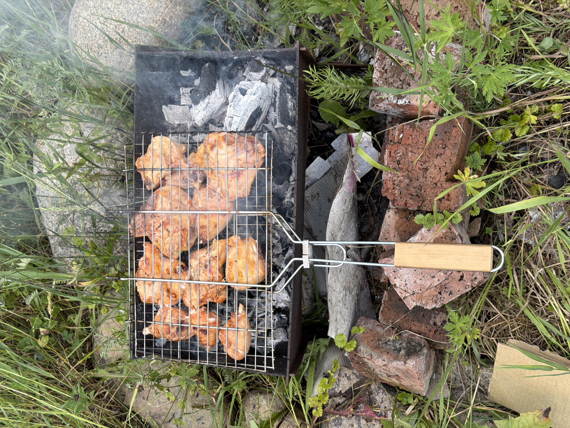 In Askat, Russia, chicken pieces are being grilled on a metal grate over hot coals in a black metal barbecue. The grill basket is filled with multiple pieces of seasoned chicken, some appearing golden brown and others lighter in color, indicating they are in various stages of cooking. Smoke is rising from the barbecue. The barbecue is placed outdoors on the ground, which is covered with grass, dirt, and scattered red bricks. Large stones are visible in the background, also surrounded by grass and vegetation. The overall environment suggests a casual outdoor setting, possibly a backyard or a picnic area.