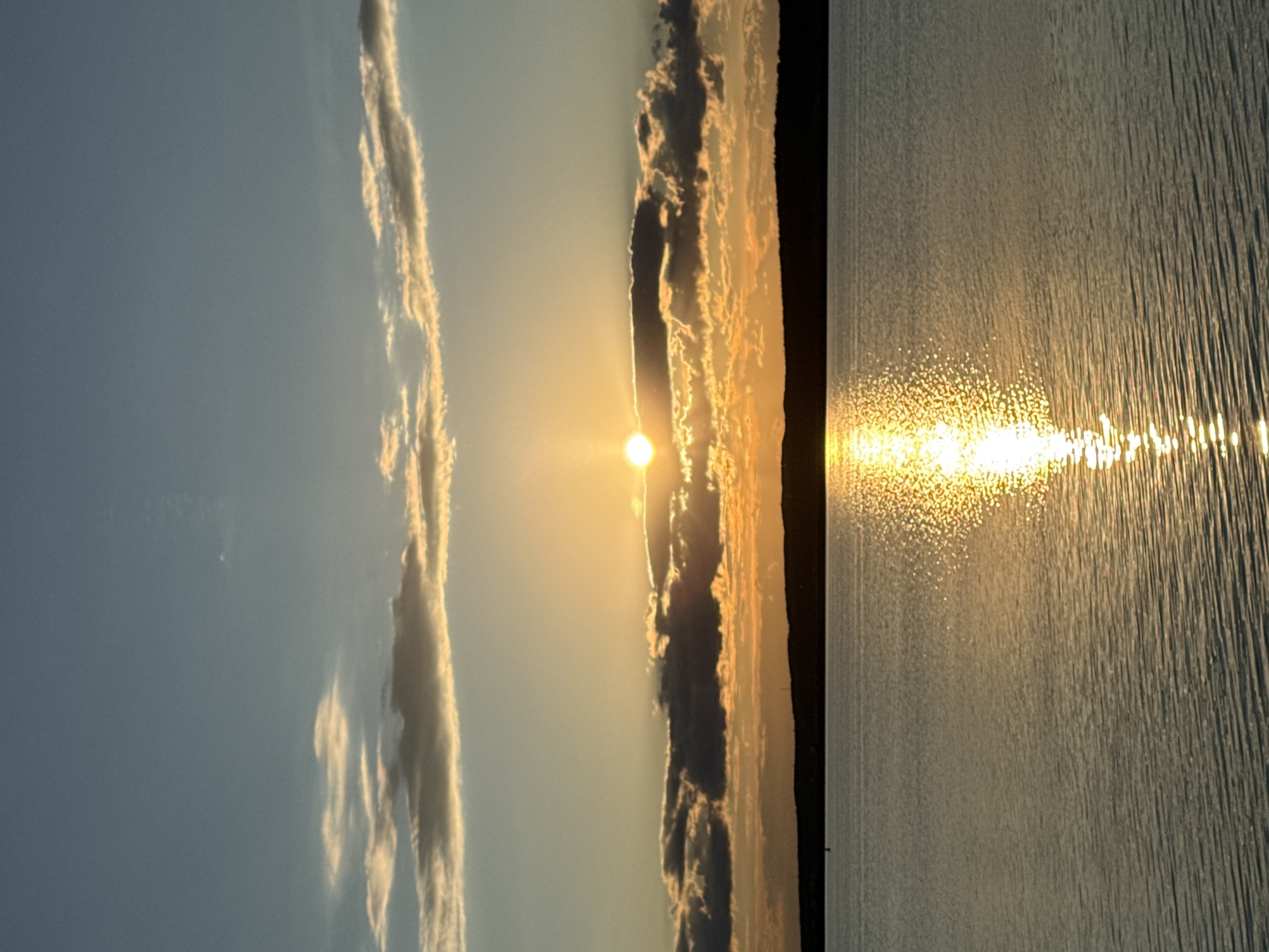 The image displays a serene landscape scene at either sunrise or sunset over a large body of water, consistent with a lake or wide river in Chaikovsky, Russia. The horizon line clearly divides the sky from the water, with a distant, silhouetted landmass visible along the horizon.
In the upper portion of the frame, the sky transitions from a soft, clear blue at the top to warm golden and orange tones closer to the horizon, bathed in the glow of the sun. Elongated clouds, some thin and wispy catching the golden light, and others appearing darker and more textured, stretch across the sky. The sun is a brilliant, intense orb positioned low near the horizon, just above the distant landmass, partially obscured by some clouds, creating a strong glare.
The lower half of the image features the surface of the water, which appears calm with subtle ripples and texture. A dazzling path of sparkling golden light, originating from the sun, stretches across the water towards the foreground, formed by numerous small reflections. The distant landmass is a dark, undefined strip on the horizon, creating a contrast against the bright sky and water.
The scene captures a tranquil moment, likely at dawn or dusk, indicated by the low position of the sun and the warm, ethereal lighting. The weather appears clear with scattered cloud formations. There are no people, man-made objects, or visible text in the image. The overall impression is one of natural beauty and peacefulness.