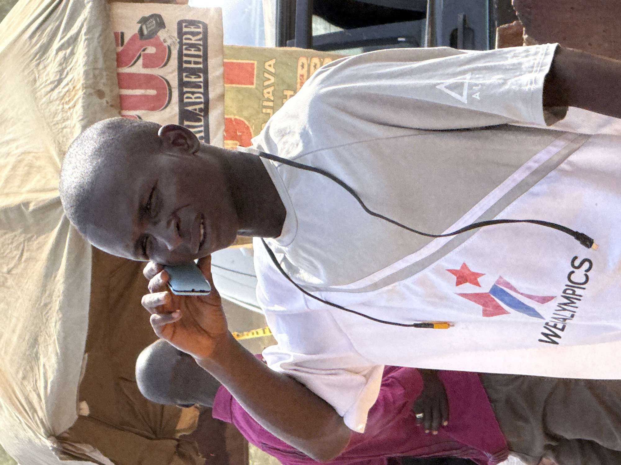 A close-up, medium-shot image depicts an African man speaking into a small mobile phone. He is wearing a white t-shirt with the word "WEALYMPICS" printed on it. The shirt also bears an emblem featuring a star, and red, white, and blue stripes. A black cable is draped around his neck. Another individual, partially visible, is wearing a purple shirt. The backdrop includes signage with legible words like "AVAILABLE HERE" and "JAVA", partially obscured by a tan-colored covering or sheet. The setting is suggested to be in Abuja, Nigeria.