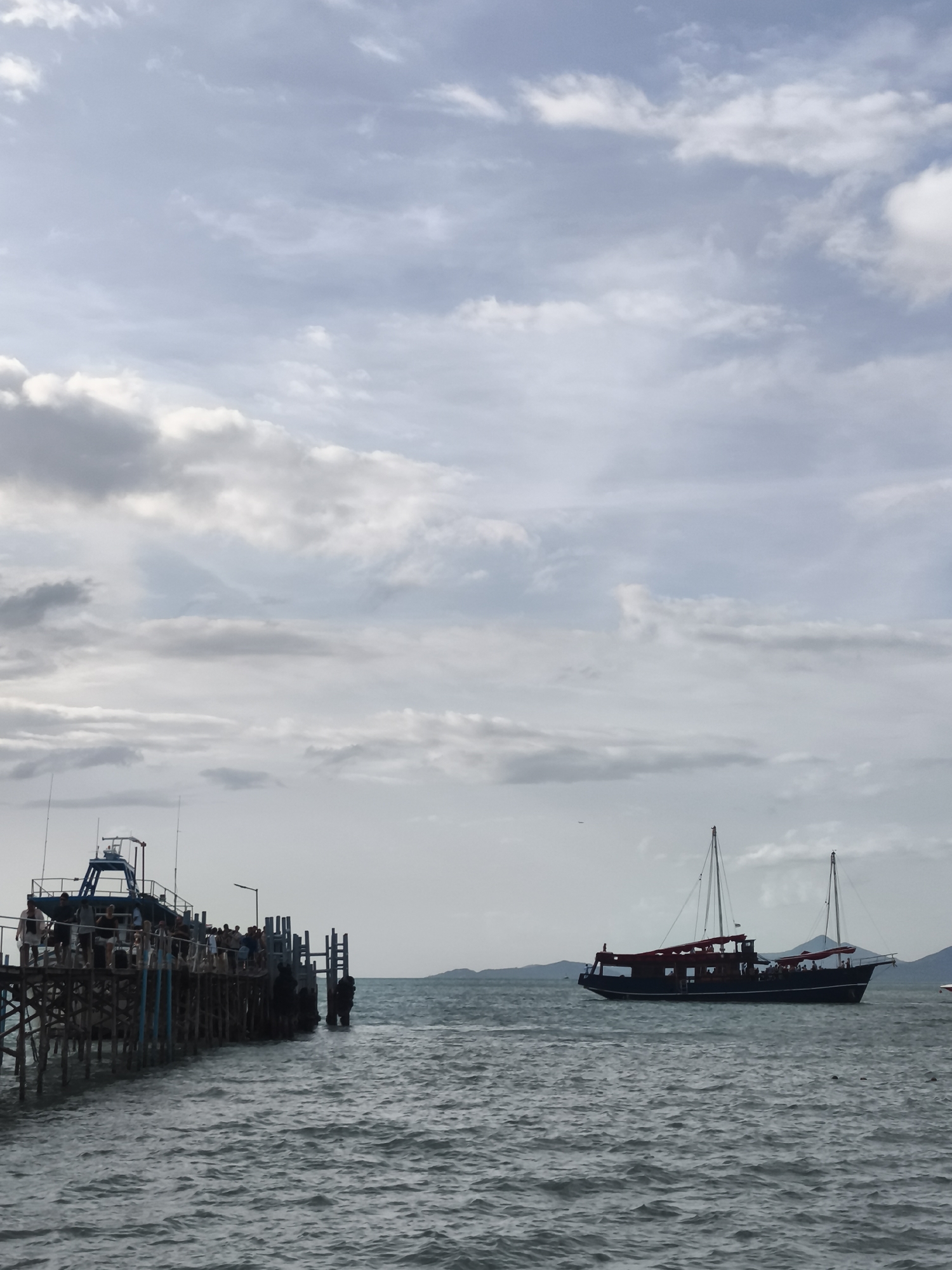 This is a photograph of a pier in Ko Samui District, Thailand. The pier is constructed of weathered wood and supported by stilts. Several people are visible on the pier, some standing and some in the process of boarding or disembarking a boat. The boat appears to be a passenger vessel. The water is choppy, indicating a moderate sea state. In the distance, a larger sailboat with red sails is visible, as well as a distant island chain. The sky is overcast with scattered clouds.