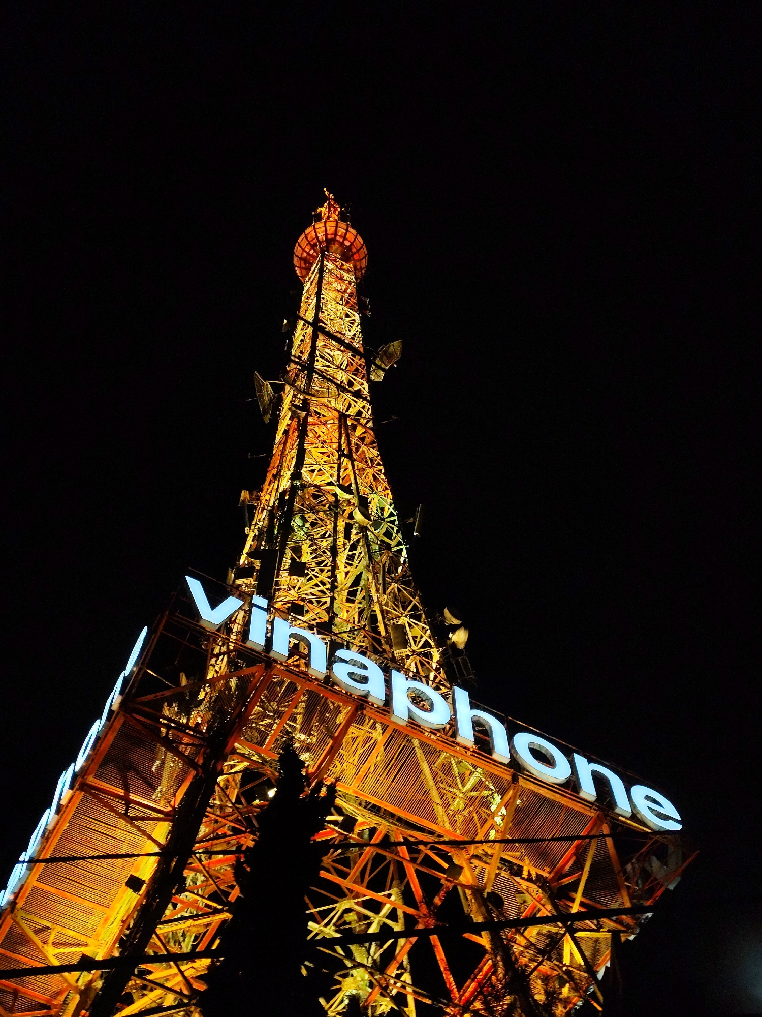 This media file features a tall transmission tower illuminated at night in Da Lat, Vietnam. The tower's structure is a lattice of metal, glowing with warm orange and yellow lights. At its base, a large sign with white, illuminated letters spells out "Vinaphone," a telecommunications company. The sign is angled, and its letters are clearly legible against the dark sky.
The scene is set at night, with the blackness of the sky providing a stark contrast to the brightly lit tower and sign. There is no discernible weather information, but the darkness suggests it is not daytime. The focus is on the man-made structure and its prominent branding. A silhouetted tree is visible in the foreground, partially obscuring the lower section of the tower, adding depth to the image. The overall impression is one of a modern, illuminated landmark against the night sky, highlighting the presence of telecommunications infrastructure.