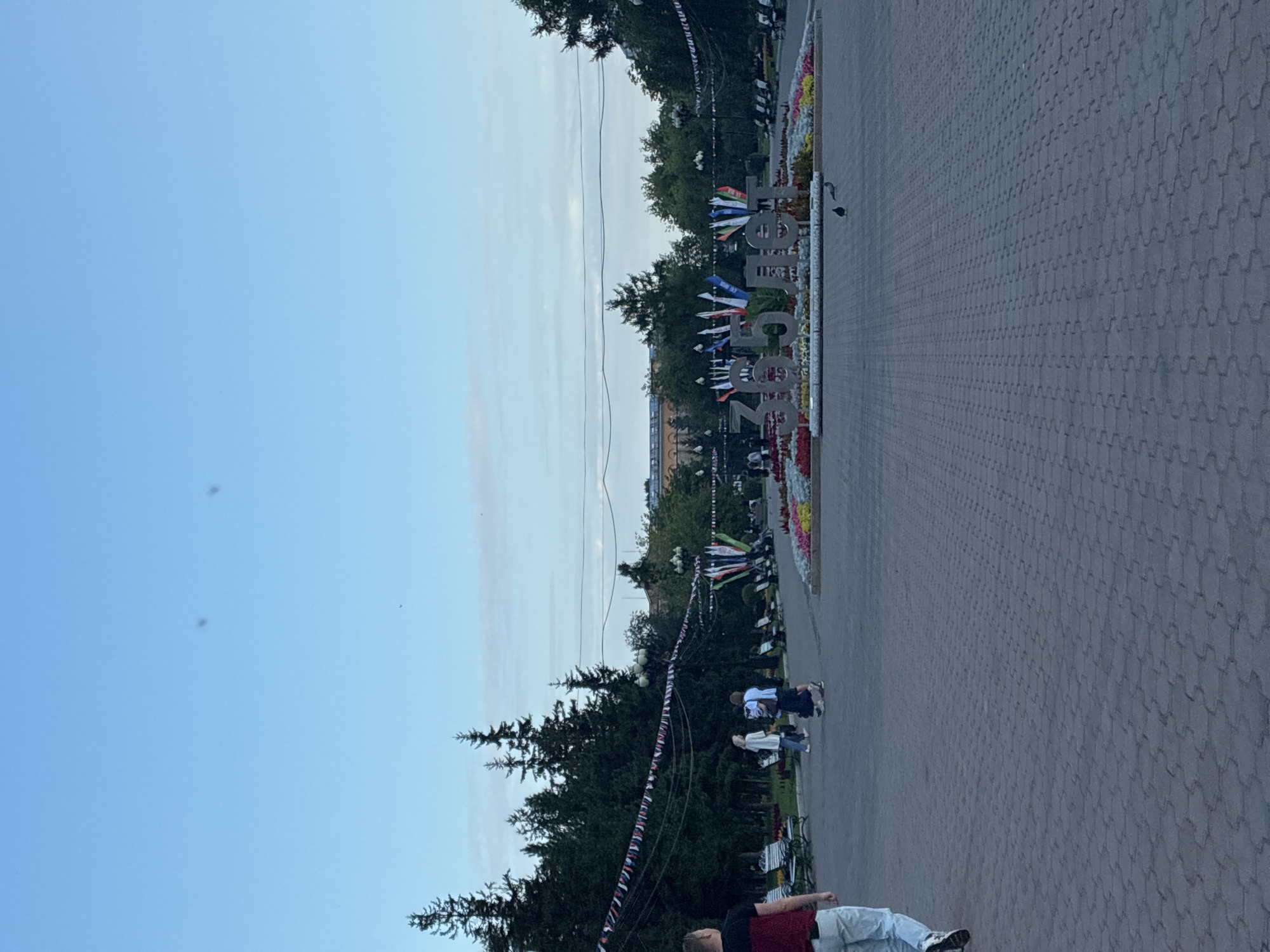 In this wide shot, a paved plaza leads towards a large, decorative display of the number "365" followed by the Cyrillic word "лет" (years), set within a flower bed. The monument is adorned with a variety of flags. To the left of the monument, a line of dark green trees stretches into the background, interspersed with lampposts and park benches. Further to the left, a man in a red and black shirt and light-colored pants walks away from the camera. To the right of the monument, the plaza continues with more trees and lights. The sky above is a pale blue with a few wispy clouds. The scene appears to be an outdoor public space, possibly a park or town square in Irkutsk, Russia, as indicated by the Cyrillic text on the monument.