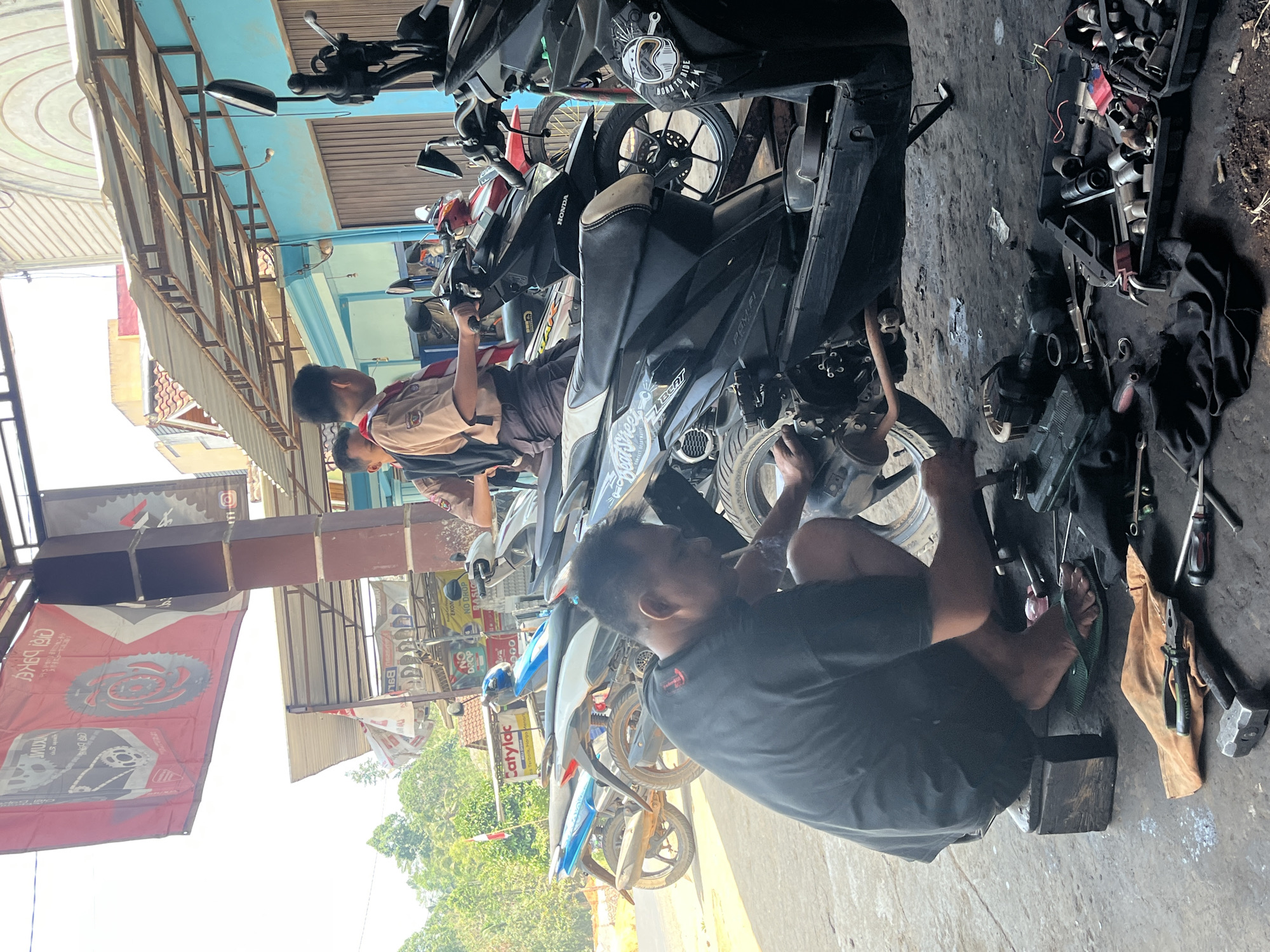 This image depicts a scene at a motorcycle repair shop in Karang Sari, Indonesia. In the foreground, a man wearing a black t-shirt and shorts is crouched down, actively working on the engine of a black motorcycle. He is holding a tool, possibly a hammer, and appears to be engaged in a repair. Various tools, including wrenches, pliers, and a toolbox filled with sockets, are scattered on the ground around him.
Behind the mechanic, several motorcycles are parked. Two young men, dressed in scout uniforms with khaki shirts and brown shorts, are standing near some of the motorcycles. One of them is holding the handlebars of a black motorcycle, while the other is looking on.
The background reveals the open-air structure of the repair shop, with a corrugated metal roof supported by posts. Brightly colored banners, one featuring images of motorcycle parts, are hanging from the ceiling. The environment suggests a typical roadside workshop in a tropical region, with lush green trees visible in the distance under a bright sky. The ground is a mix of concrete and dirt, typical of such outdoor establishments.
