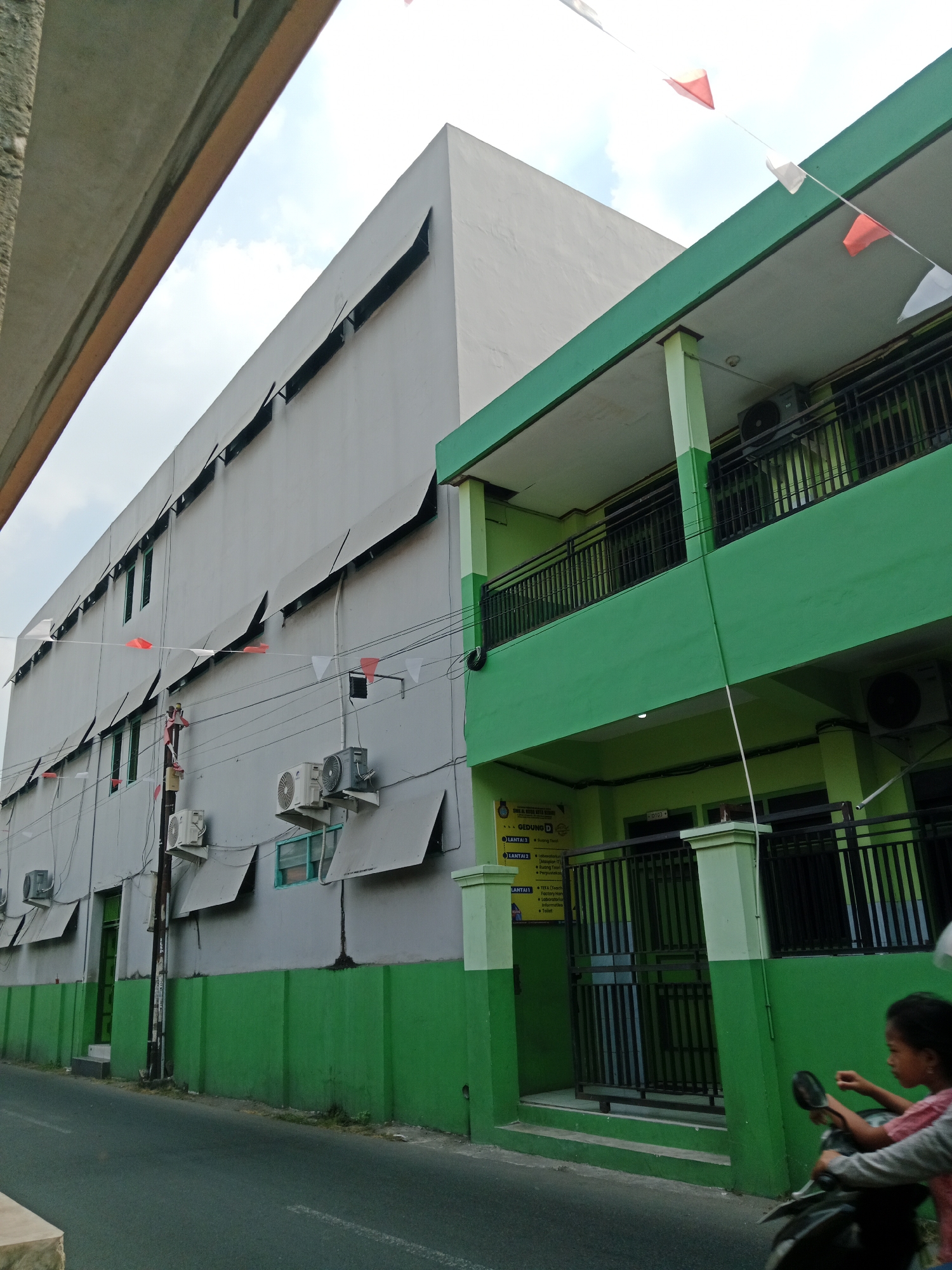 The image shows a street view of a two-story building with a large, gray, multi-story structure adjacent to it. The building on the right is painted green and has balconies with black metal railings on both levels. Small red and white triangular flags are strung across the top of the scene, suggesting a festive occasion. Air conditioning units are visible on the exterior walls of both buildings. A portion of a concrete overhead structure is visible on the left side of the image. In the lower right quadrant, a person is riding a motorcycle, partially visible, with the handlebars and a portion of the person's upper body in view. The street is paved and appears to be in a residential or commercial area. The overall scene depicts a typical urban environment in Kediri City, Indonesia, characterized by its architecture and the presence of everyday activities.