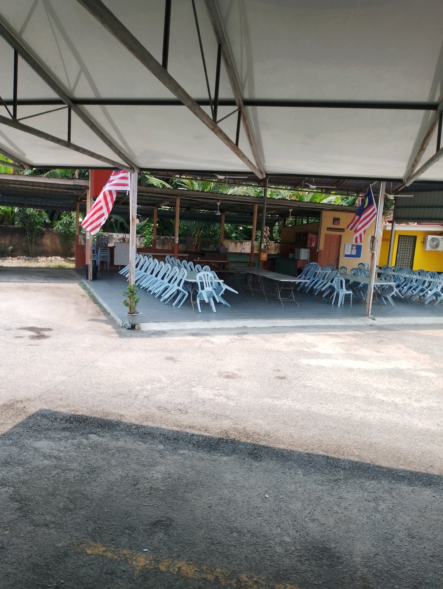The image depicts an outdoor area with a covered seating arrangement, likely for events or dining, located in Pulau Tiga, Malaysia. The scene is dominated by rows of empty, light blue plastic chairs arranged around foldable tables under a white canopy supported by metal beams. The Malaysian flag is prominently displayed on a flagpole in the foreground, adding a patriotic element. Behind the seating area, a building with yellow and muted brown walls is visible, featuring windows, a door, and an air conditioning unit. Lush green foliage and trees are present in the background, suggesting a tropical environment. The ground is paved with asphalt, with some areas appearing lighter in color, possibly indicating concrete or a different type of paving. The overall impression is that of a prepared venue, awaiting occupants for an outdoor gathering.