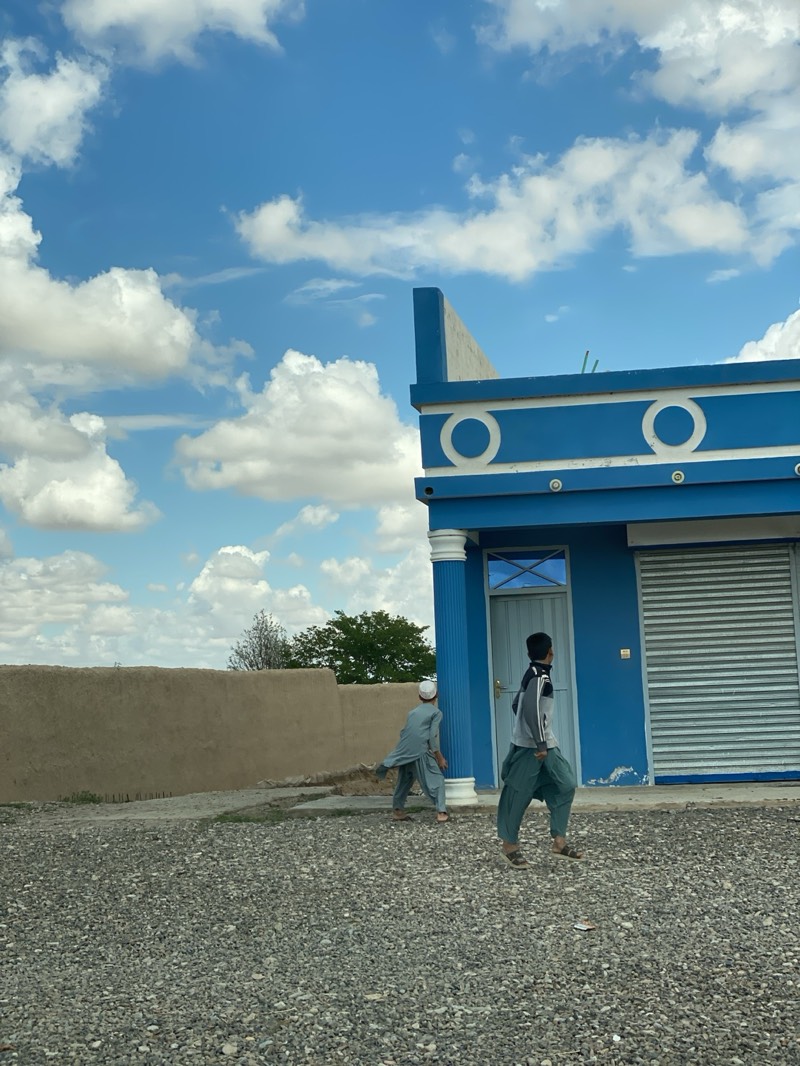 The image, likely captured in Mandisar, Afghanistan, depicts an outdoor scene on a bright, sunny day with scattered cumulus clouds in a blue sky. In the foreground, a gravel-covered area dominates. Two young males are present. One boy walks toward the viewer, wearing loose-fitting green pants and sandals, and a gray and white long-sleeved shirt. Another boy, further back, is facing away, wearing similar loose-fitting light gray clothing and a white skullcap. To the right is a single-story blue building, possibly a store or shop, with a closed metal roll-up door and a blue door framed by white faux pillars. To the left is an aged, tan-colored wall extending into the background. Beyond the wall and between the two boys is a patch of green trees or shrubbery. The overall environment suggests a rural or semi-rural setting within Mandisar.