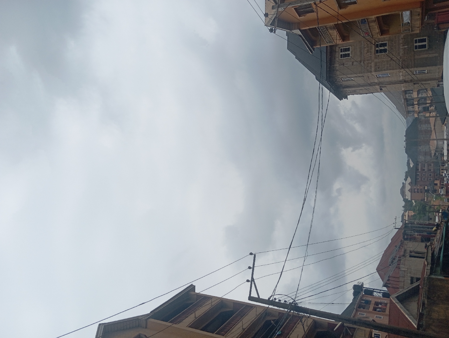 This image presents a view looking upwards towards a heavily overcast sky in an urban environment, specifically Nkpor, Nigeria. The vast majority of the frame is dominated by dense, grey clouds, indicating a very cloudy day, possibly preceding or during rainfall.
Multi-story buildings frame the scene on the left, right, and bottom sections. These structures appear to be a mix of residential and commercial properties, exhibiting various architectural styles and states of repair. Some buildings have light-colored painted facades, while others show exposed concrete or more weathered finishes, with multiple windows and balconies visible. The density of these buildings suggests a well-developed urban or semi-urban area.
Numerous power lines traverse the sky, strung between utility poles that are visible in the midground and foreground, particularly on the lower right where a prominent pole with multiple cables is seen. On the rooftop of a building in the lower right, a black plastic water storage tank is visible, a common feature in the region. In the distant background, a horizon line of more buildings and potentially undulating terrain is discernible beneath the heavy cloud cover.
The absence of direct sunlight and the uniform grey sky contribute to a somber or muted atmosphere. There are no people visible, nor any specific events or activities taking place. No discernible text is visible on any of the structures or objects. The image captures a quiet moment of an urban landscape under a perpetually cloudy sky.