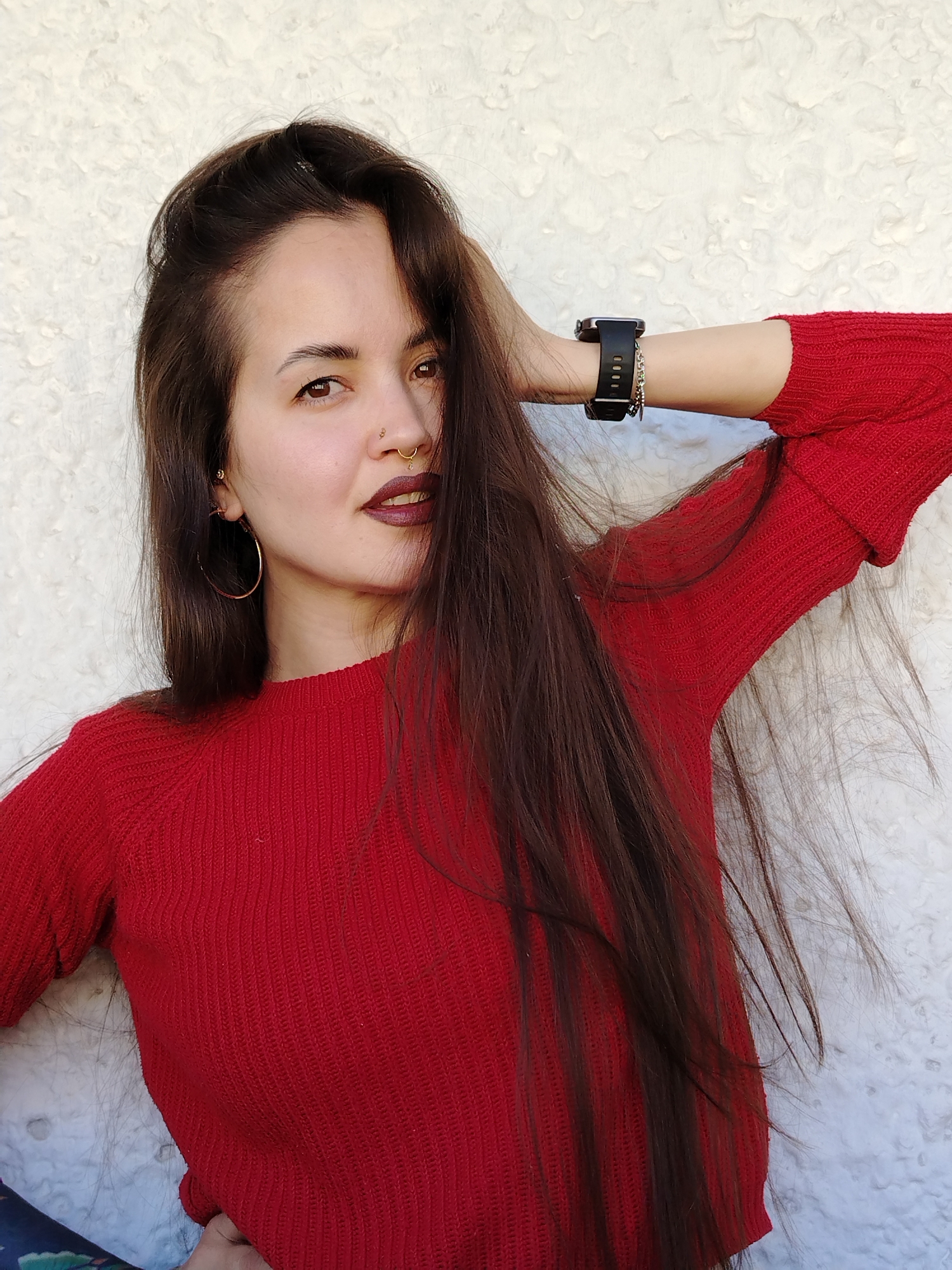A woman poses indoors against a white textured wall. She is wearing a red sweater, has long brown hair, and wears jewelry, including a nose ring, hoop earrings, and a black smartwatch on her left wrist. Her right hand is raised and resting behind her head. Part of a patterned object is visible at the bottom left corner of the frame. There is no definitive evidence to pinpoint this image's location in Noviy, Russia.