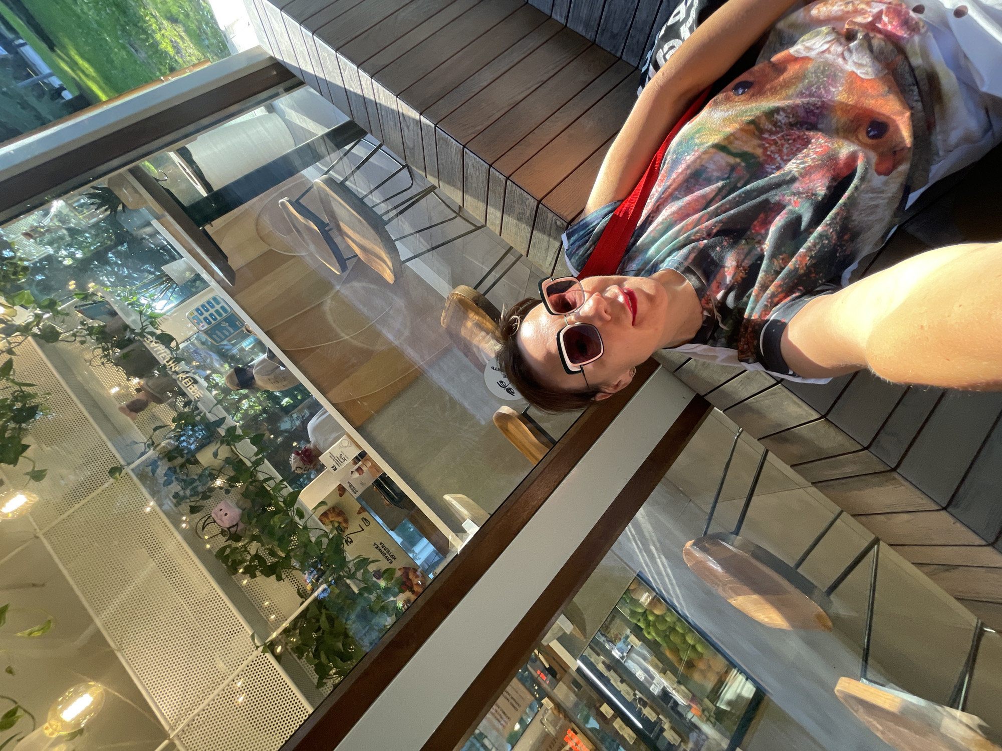 A woman is taking a selfie while lying on a wooden bench outdoors, likely in Sochi, Russia. She is wearing a colorful t-shirt with a large print of a fox's face and sunglasses. The setting appears to be a cafe or a public area with large glass windows. Through the windows, plants and shelves with products are visible, suggesting an indoor space. Sunlight streams onto the scene, indicating it's daytime. The woman's expression, with a slight smile, conveys a relaxed and happy mood. The wooden bench is modern in design, and the overall ambiance suggests a casual, sunny day.