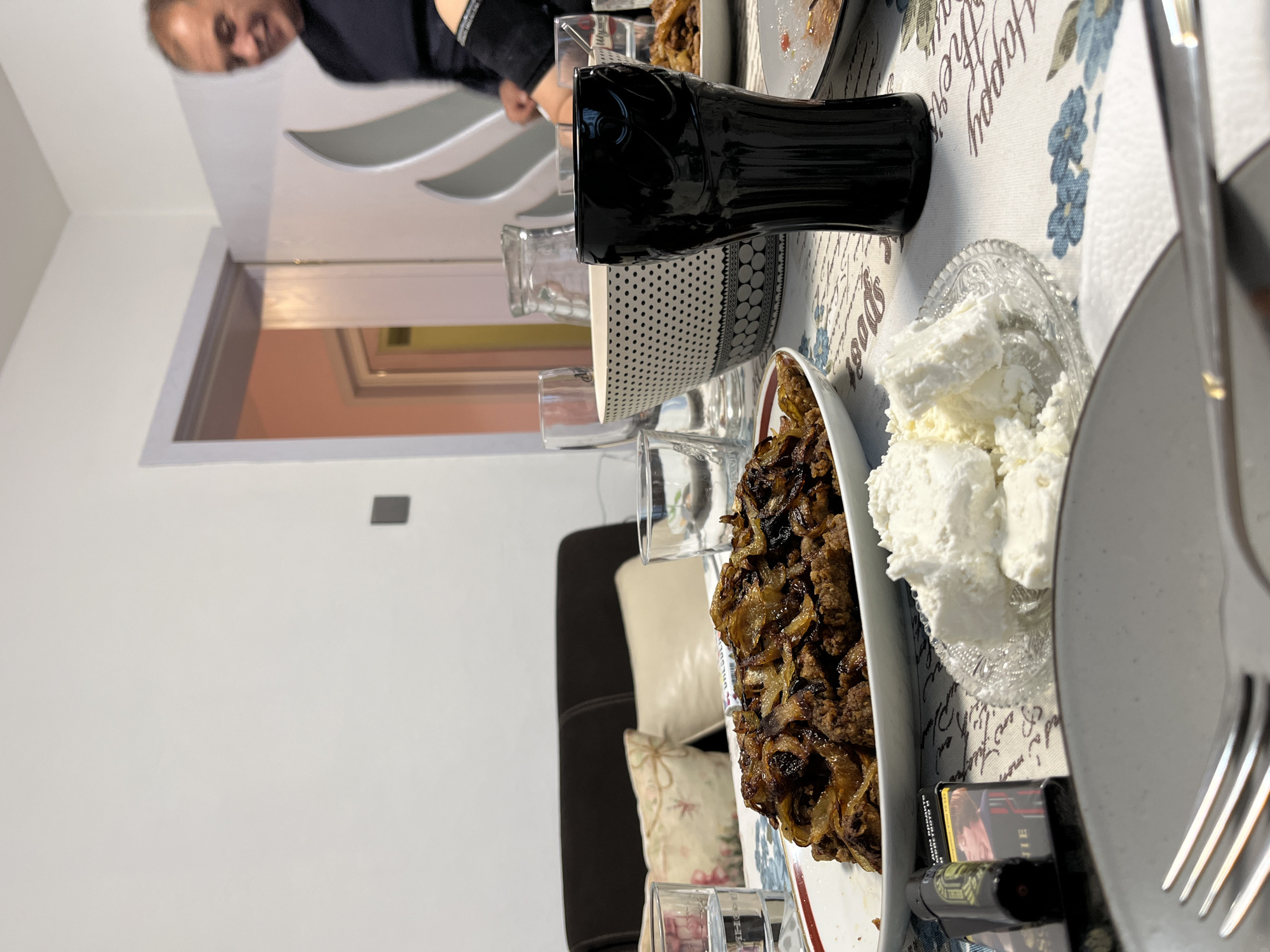A table is set for a meal with food and beverages. Dishes on the table include caramelized onions, fresh white cheese or cream cheese, and what appears to be a Coca-Cola bottle. Cutlery and plates are present, suggesting a meal in progress or about to begin. A man is visible partially in the background, out of focus. The tablecloth has a floral design and text, which is illegible. The location is not discernable from the image. Karavelovo, Bulgaria cannot be determined.