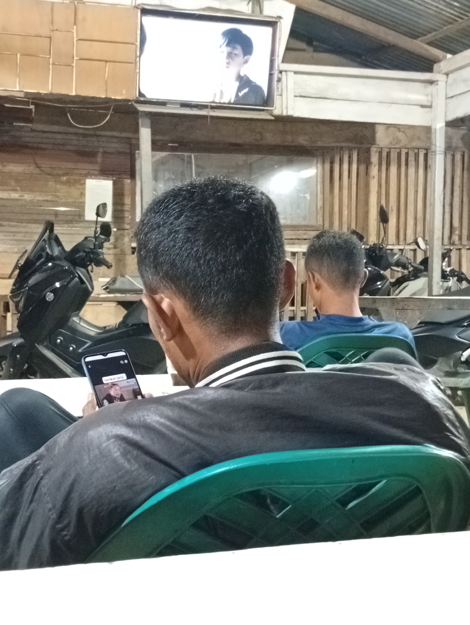 The image depicts an indoor scene with several individuals. In the foreground, a person is seated, facing away from the camera, and holding a smartphone. The phone screen displays a video of a person, and text is visible below it. To the left, a black motorcycle is parked. Behind the seated individuals, another person is visible from the back, also seated. Further in the background, a television screen shows an image of a person. The environment appears to be a covered area with wooden walls and a corrugated metal roof. The presence of motorcycles and the general aesthetic suggest this location could be a roadside stall or a workshop in Lhokseumawe, Indonesia.