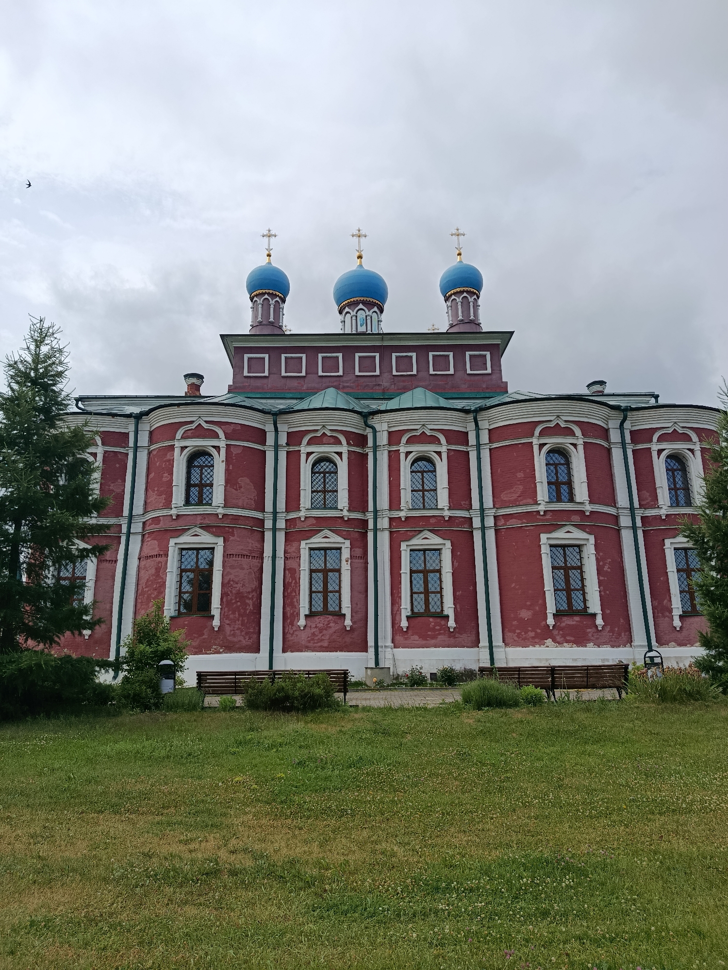 The image shows the Assumption Cathedral in Radovitsy, Russia. The cathedral is a large, red brick building with white trim and a green roof. It has three blue domes topped with golden crosses. The building is surrounded by a grassy lawn with several benches. There are trees on either side of the cathedral. The sky is cloudy. The image is taken from a low angle, looking up at the cathedral.