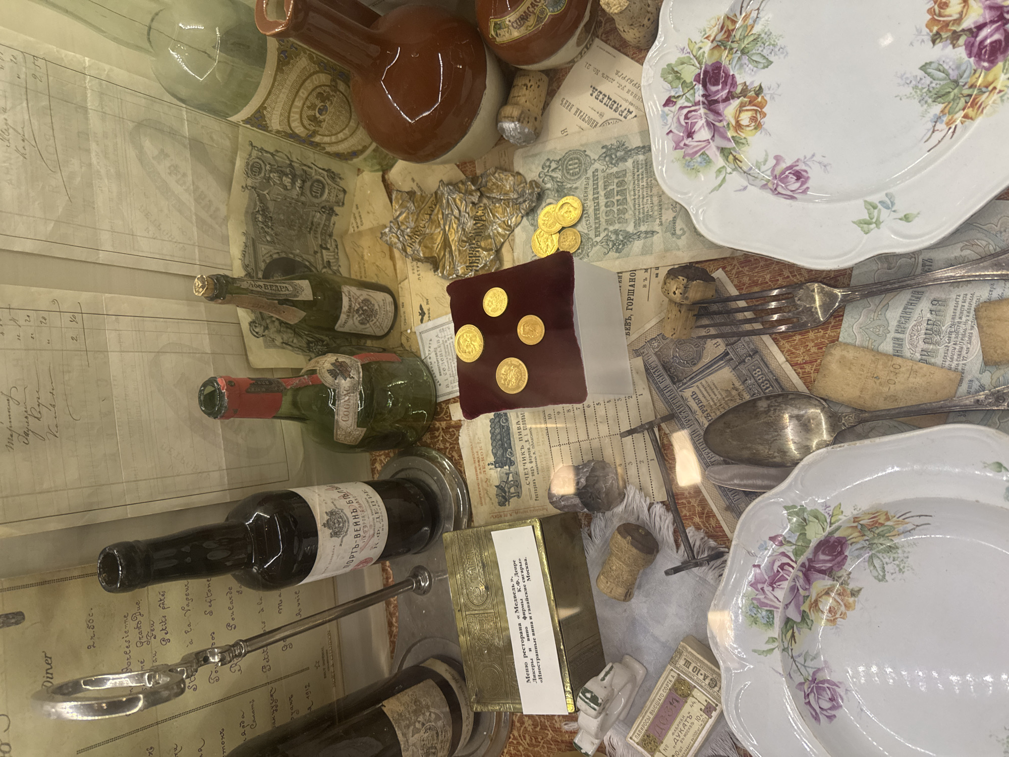 The image presents a display case exhibiting an assortment of antique objects. There are several bottles of wine and liqueur, along with vintage plates and silverware. The backdrop features old documents, possibly receipts or menus, and currency notes from the era. A burgundy cushion displays gold coins. A menu from the "Medved" restaurant, known for its liquors, wines, and cigars, is also visible. Corks, a silver fork, and cigarette boxes are scattered throughout the display. This setting evokes a historical ambiance and suggests a connection to the city of Moscow, Russia through the menu card and wine labels. There is no indication that this display is located in Saint Petersburg.