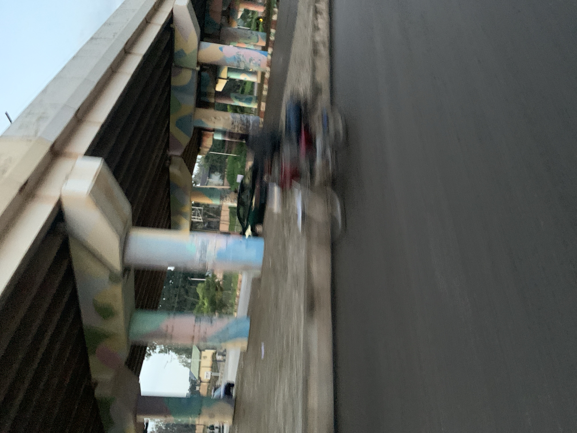This image depicts a blurred scene of a highway overpass in Jos, Nigeria. The concrete pillars supporting the overpass are decorated with colorful, abstract graffiti in shades of pink, green, blue, and yellow. Below the overpass, on the road surface, a motorcycle with a rider is captured in motion, appearing as a blur of red and dark colors. On the raised walkway to the right of the motorcycle, another blurry figure is visible. Further in the background, a dark-colored car is parked near a building with a light-colored roof, partially obscured by trees and the overpass structure. The sky appears overcast.