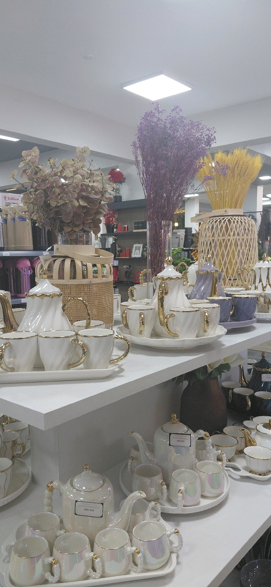 The image captures a retail display shelf filled with various tea sets. In the foreground, a white tea set with gold accents is prominent, featuring a teapot and several cups on a tray. To the right, another tea set in white with gold trim is visible. Further back, there are more tea sets in white, some with iridescent finishes, and some in a lavender hue. The background shows shelves with decorative items, including dried flowers in vases and woven baskets. The overall setting appears to be an indoor store, likely a home goods or decor shop. The location is in Encarnacion, Paraguay, as indicated by the price tag on one of the tea sets, which shows "385.000", a typical currency format for Paraguay.