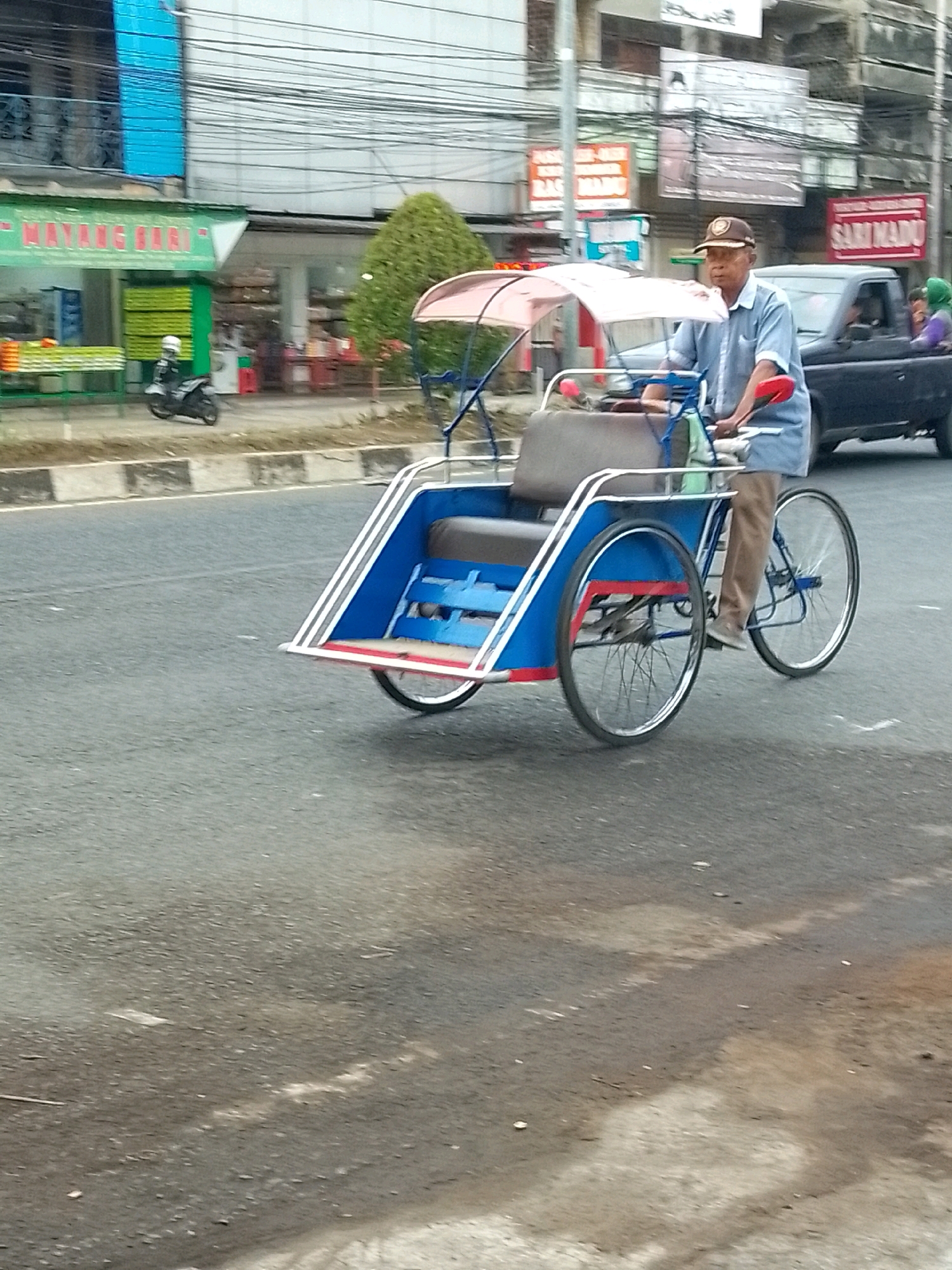 In a street scene in Unknown, Unknown, a man wearing a light blue collared shirt, brown pants, and a brown baseball cap is operating a blue and white pedal rickshaw. The rickshaw has a large, curved canopy and a passenger seat. The man is pedaling and appears to be driving the rickshaw down the asphalt road. In the background, a black pickup truck is visible, and behind it, several buildings line the street. A green awning with white lettering is prominent on one of the buildings. A parked motorcycle with a white rider's helmet is also visible on the sidewalk. The overall environment suggests an urban setting with commercial activity.