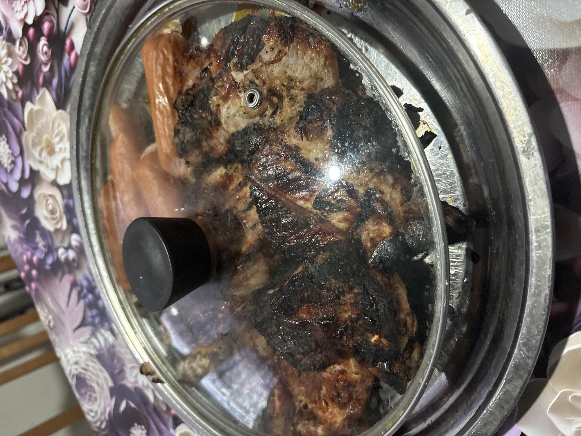 A close-up, overhead shot captures a lidded metal container filled with cooked food, resting on a patterned surface. The container, possibly a pot or a slow cooker insert, holds what appears to be heavily grilled or roasted meat, characterized by dark, charred patches and lighter brown sections. Alongside the main meat, several elongated, lighter-colored sausage-like items are visible.
A clear glass lid covers the food, featuring a prominent black handle/knob in the lower left portion and a small metal vent ring near the center. The lid shows some condensation or steam, suggesting the food beneath is warm. The metal rim of the container is visible around the lid, exhibiting some wear and minor blemishes.
The container is placed on a tablecloth or mat adorned with a floral design, featuring white and purple blossoms against a darker background. The overall scene suggests an indoor setting, likely a home kitchen or dining area where prepared food is being kept warm or presented. No individuals are visible, nor is any text.