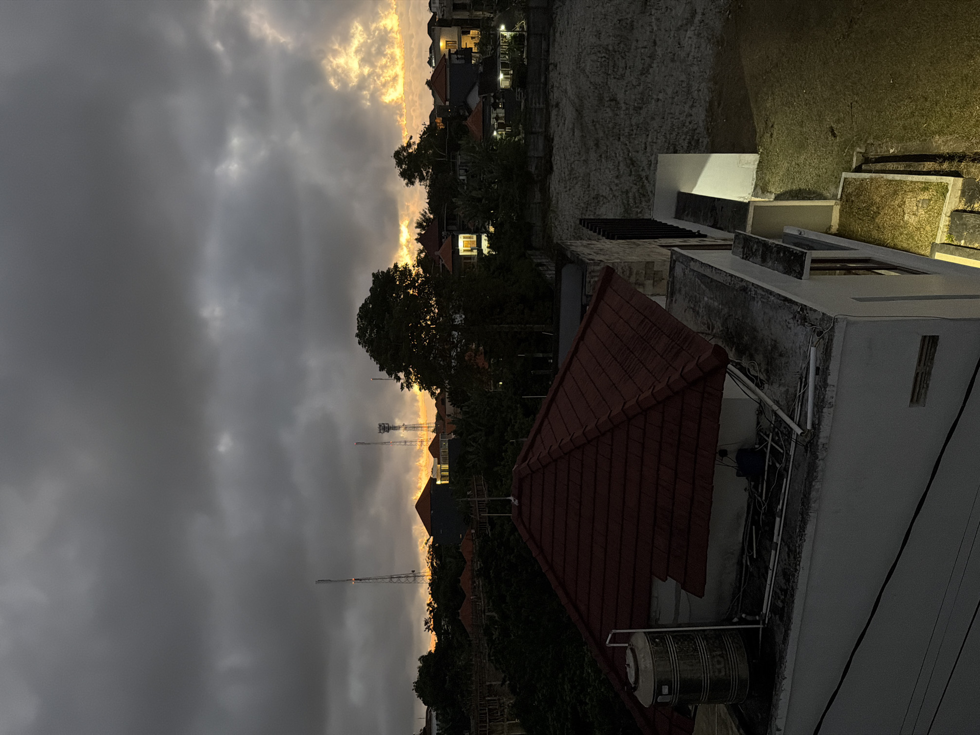 The image, likely taken in Kuta Selatan, Indonesia, shows a dusk or dawn scene viewed from a rooftop. The sky is dominated by heavy, overcast clouds, with a sliver of golden light visible near the horizon. In the midground, a mix of residential buildings and dense trees line the landscape, punctuated by the silhouettes of telecommunication towers. The foreground features the rooftops of adjacent structures, including a distinctive red-tiled roof and various utility installations such as a water tank and electrical wiring. The overall atmosphere is one of subdued light and urban density.