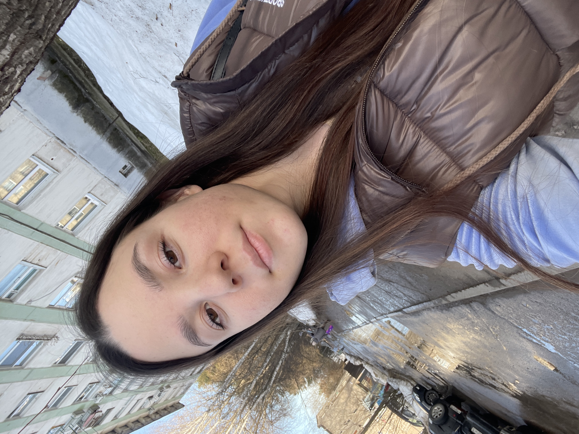 A medium shot depicts a young woman wearing a brown puffy vest and a light blue shirt, taking a selfie outdoors in what appears to be the city of Chaikovsky, Russia. Her face is the most prominent element, with brown hair partially obscuring the upper right of the frame. Behind her, the environment features urban elements including multi-story apartment buildings with light-colored facades, trees without leaves, and a street partially covered with melting snow and standing water. Cars are parked along the street. A distant figure of a person can be seen walking. The overall tone suggests a cold, early spring day.