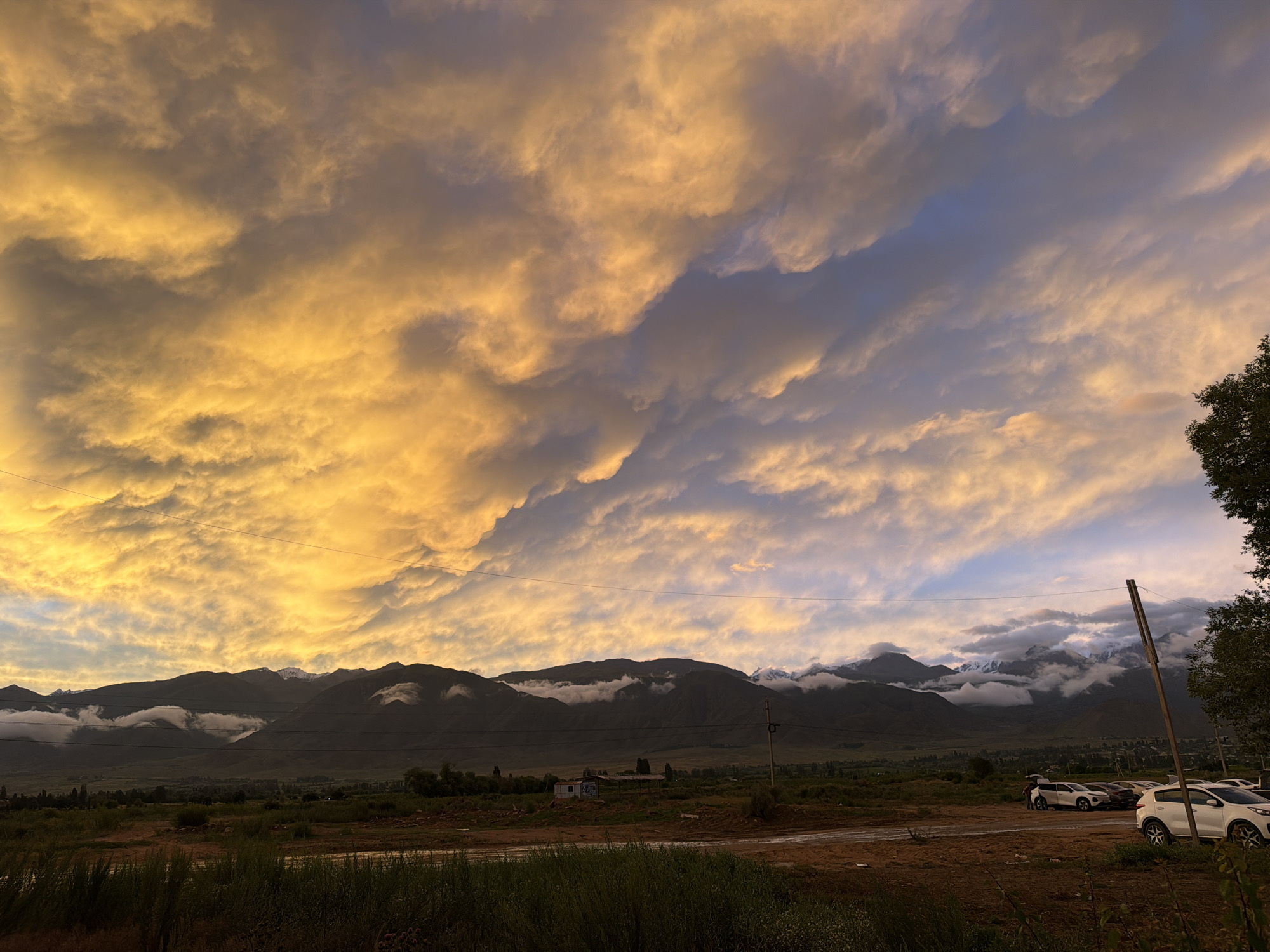 This image captures a dramatic sunset over the mountains near Çolpon-Ata, Kyrgyzstan. The sky is dominated by heavy, textured clouds illuminated in vibrant shades of orange and yellow, contrasting with patches of deep blue and purple. Below the striking sky, a range of dark, imposing mountains stretches across the horizon, some peaks capped with snow or partially obscured by low-hanging clouds.
In the foreground, a flat, open area of dirt and dry grass appears to be a parking lot. Several white SUVs are parked here, with one car door open and a person visible inside, suggesting a brief stop or departure. A single wooden utility pole stands prominently on the right side of the frame. Further back, a small, rudimentary building with a tin roof is visible in the middle ground, amidst sparse vegetation and a few trees. The overall environment suggests a rural or semi-urban setting on the outskirts of a town. The low light conditions indicate either dawn or dusk.