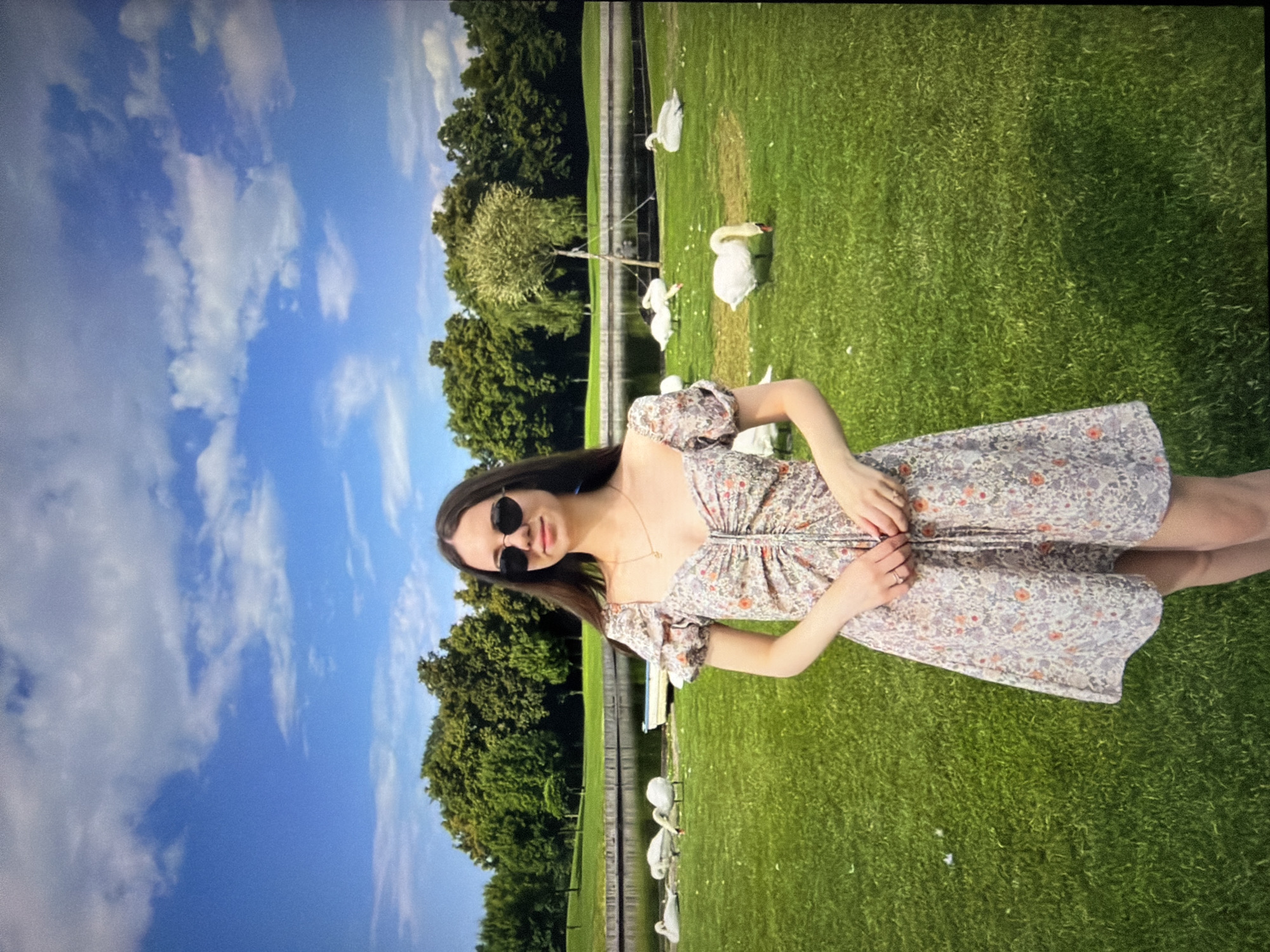 In an outdoor setting with a lush green lawn and a body of water in the background, a young woman stands and poses for the camera. She is wearing a floral print dress and sunglasses. Several swans are scattered around the lawn and near the water's edge. The sky above is blue with scattered clouds. The location is identified as Kyiv, Ukraine.