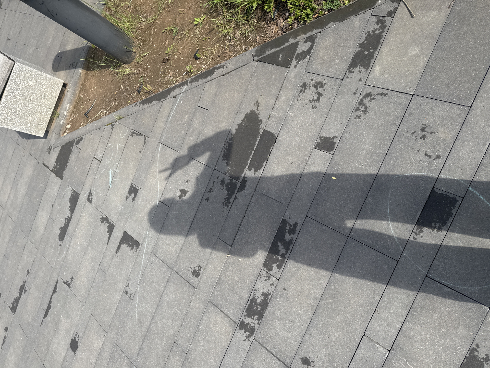 The image shows a shadow of a person standing on a paved walkway in Izhevsk, Russia. The walkway is made of dark gray rectangular pavers, some of which are wet, indicating recent rain or watering. The shadow is cast by the sun, which is positioned to the left and slightly behind the viewer. The person in the shadow appears to be holding their left arm up, with their fingers forming a peace sign. There are some faint white chalk marks on the pavers, possibly drawings or game markings. To the right of the walkway, there is a patch of dirt with some sparse grass and a dark gray cylindrical post. Further in the background, a light gray concrete bench or planter box is visible. The overall scene suggests an outdoor public space.