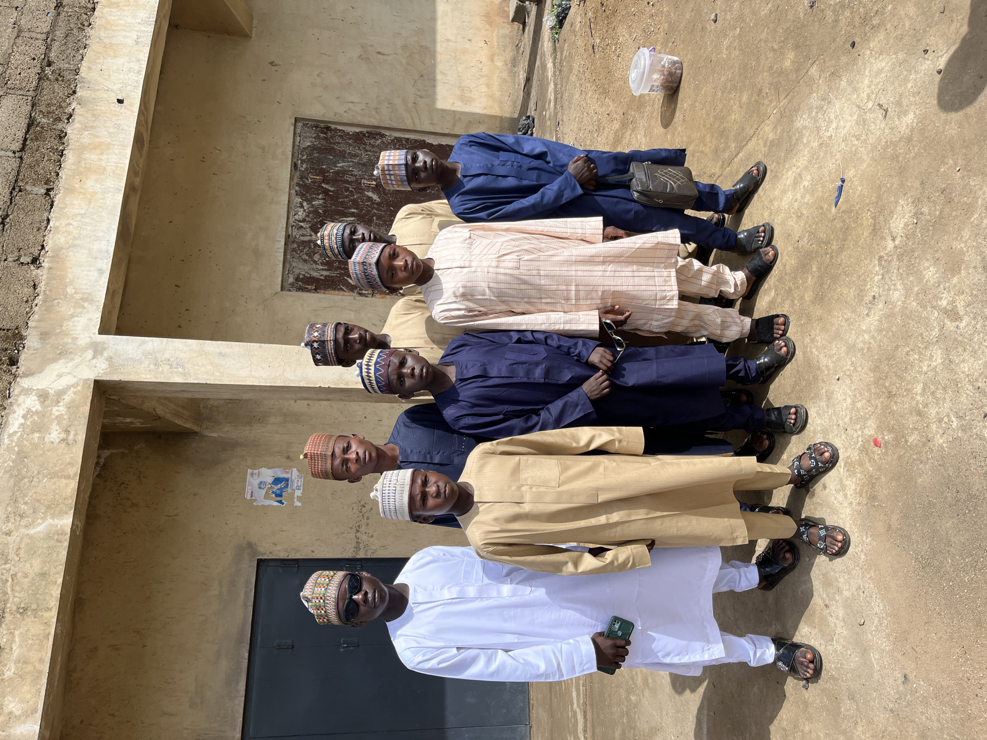 A group of seven people stand together in Naraguta Hausawa, Nigeria. They wear clothing typical of Hausa culture, including embroidered caps. The person at the front is wearing sunglasses and holding a phone. They stand in front of a weathered building. A political poster is partially visible on the wall. A plastic container rests on the ground near the group.
