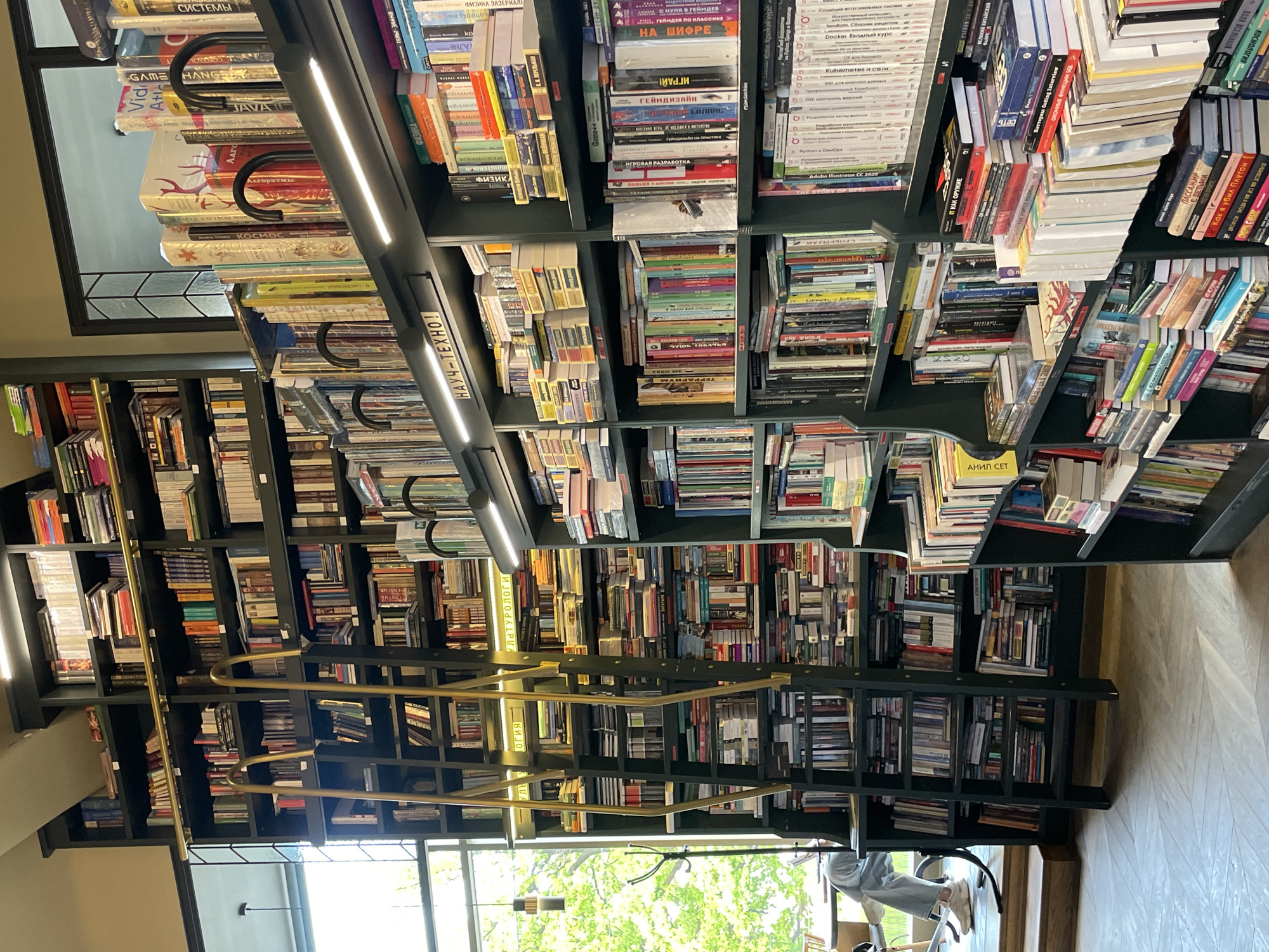 The image depicts a bookstore in Saint Petersburg, Russia. The shelves are filled with numerous books, showcasing a diverse range of titles in both Russian and English. A rolling ladder on a gold track provides access to the upper shelves. The bookstore is illuminated by bright ceiling lights. A person is seated on a chair near a window, likely browsing through books. The scene has a relaxed and inviting atmosphere, characteristic of a literary space.