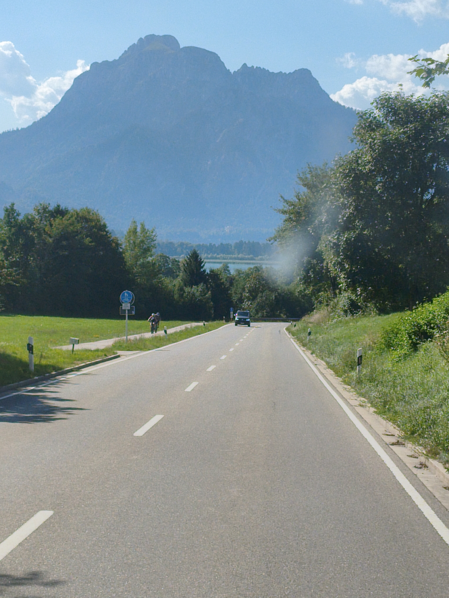 The image depicts a paved road winding through a green landscape with a large mountain in the background. The mountain, likely the Säuling, is a prominent feature of the Füssen, Germany, region. A white car is visible on the road, approaching the viewer. To the left of the road, there is a paved path with two cyclists. A blue and white traffic sign with a bicycle symbol is posted near the path. Trees and grassy fields line the road, and a body of water, possibly a lake, is visible in the distance beyond the trees. The sky is blue with scattered white clouds. The overall scene suggests a scenic drive or cycling route in the Bavarian Alps.