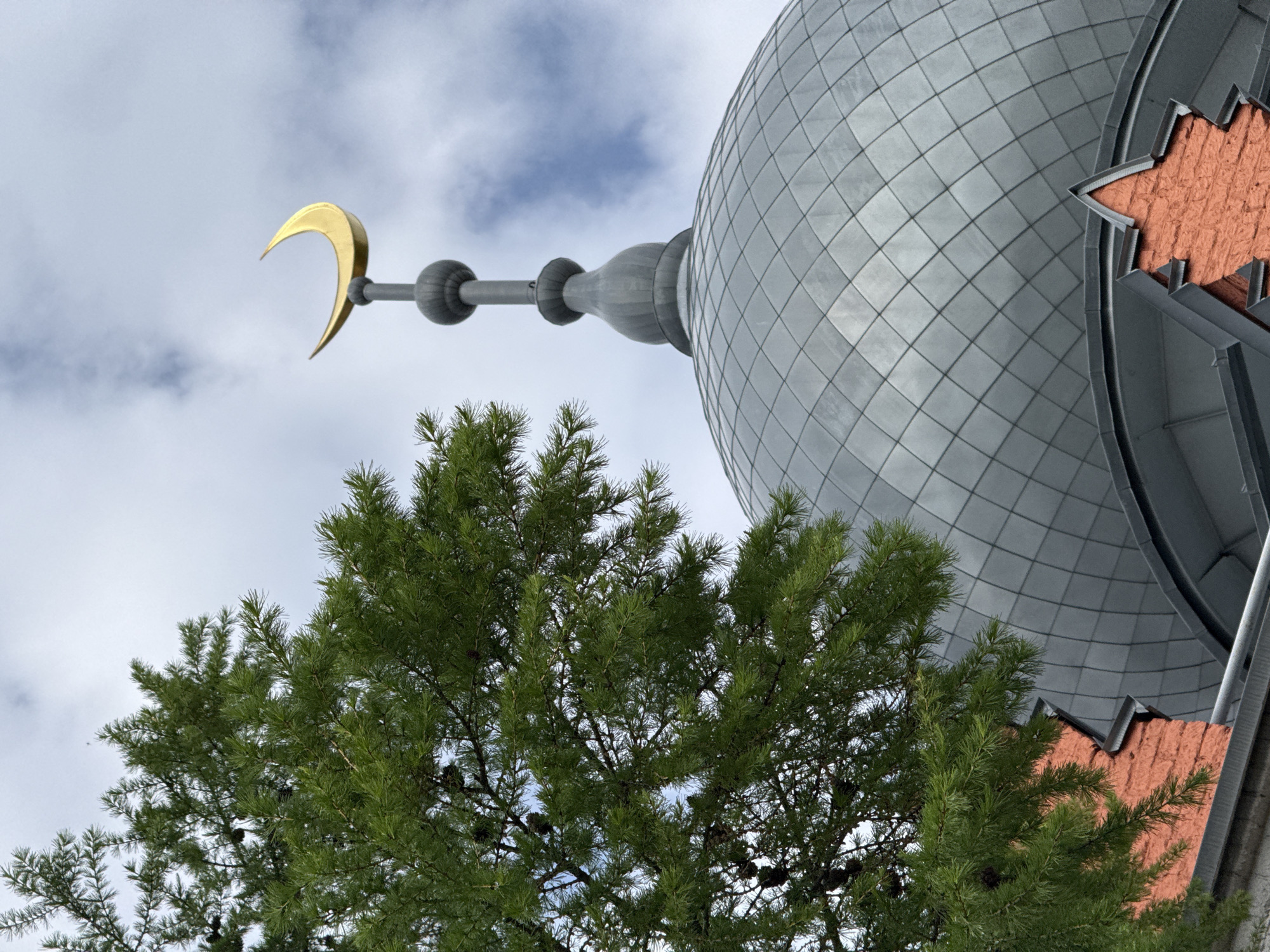 A low-angle shot captures a portion of the Nizhny Novgorod Mosque in Russia. A golden crescent moon sits atop a long, gray pole extending from the gray, tiled dome of the mosque. The dome has red, star-shaped accents. A large, green tree partially obscures the view of the structure. The background consists of a cloudy sky.