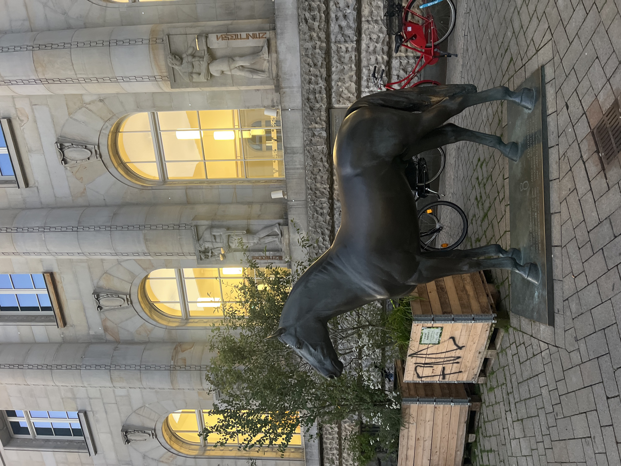 In Oldenburg, Germany, a bronze sculpture of a horse stands on a paved sidewalk in front of a stone building. The building has several large arched windows with yellow light emanating from within, and decorative sculptures of figures on either side of the windows. Two wooden planters filled with greenery are to the left of the horse statue. A red bicycle with a basket is parked to the right of the horse, and another bicycle is partially visible behind the horse. The horse is mounted on a bronze plaque with engraved text. The scene appears to be an outdoor public space, possibly a plaza or a street.