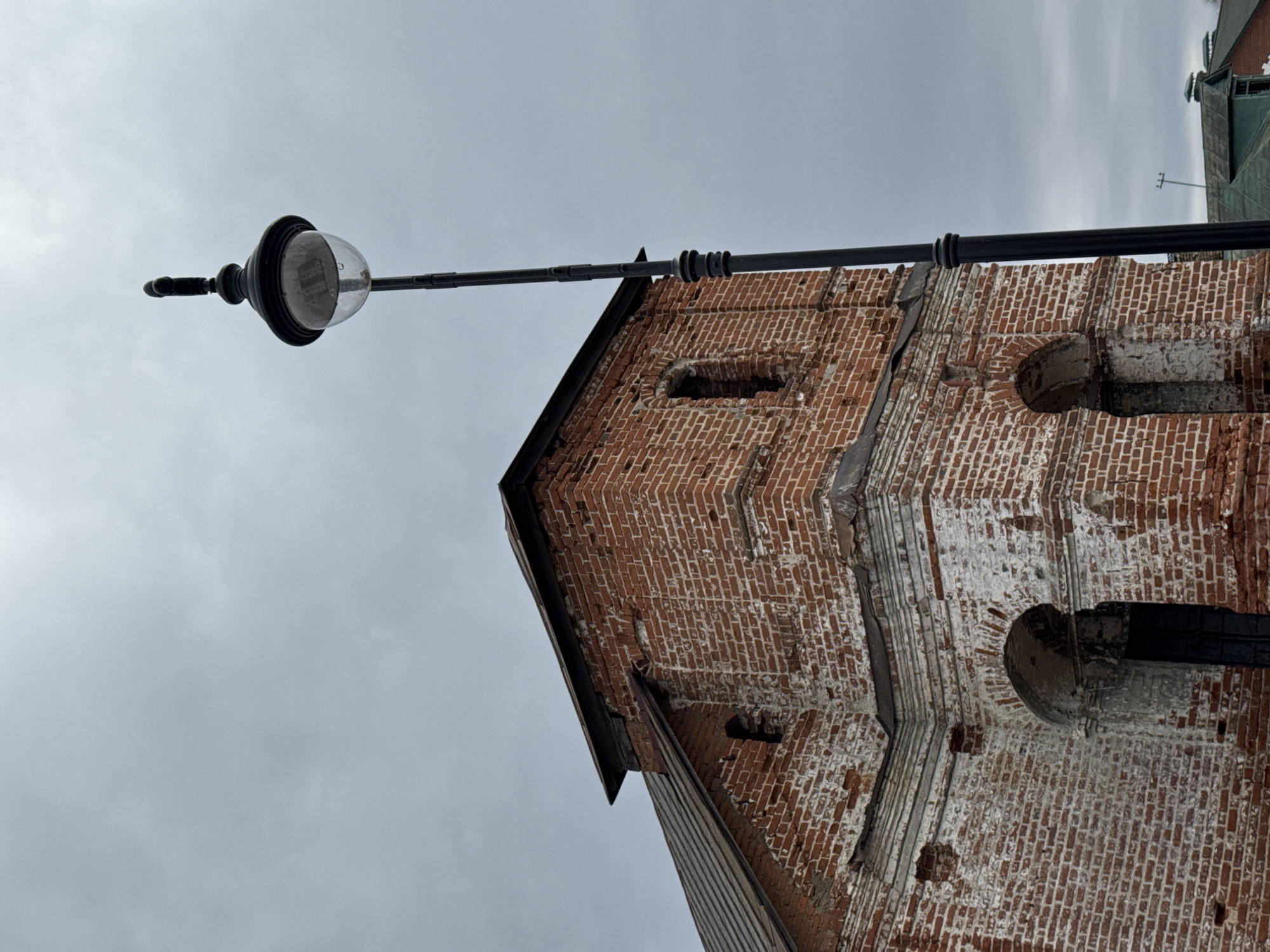 The image shows the exterior of a brick building in Ново-Никольское, Russia. The building has a weathered, aged appearance, with faded white paint visible on some of the brickwork. There are architectural details such as arches and rectangular indents in the brick. A black metal street lamp extends over the building, silhouetted against a cloudy sky. The image is taken from a low angle, looking up at the building and the lamp.