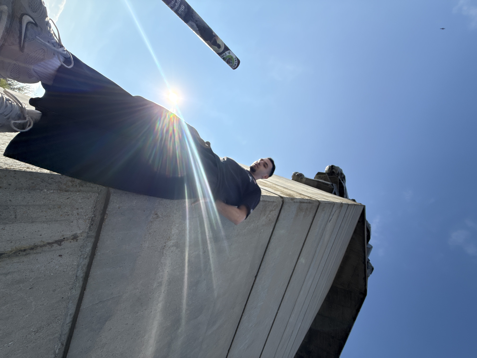 A low-angle shot shows a person lying on a concrete structure under a clear blue sky with the sun shining directly overhead. The person is wearing a dark t-shirt and dark pants or shorts. The person appears to be wearing grey sneakers. Above the person's head is a long, cylindrical object with writing on it. The structure is made of concrete slabs. The location is the Founders of the Bulgarian State Monument in Shumen, Bulgaria. A small dark object is also visible in the sky.