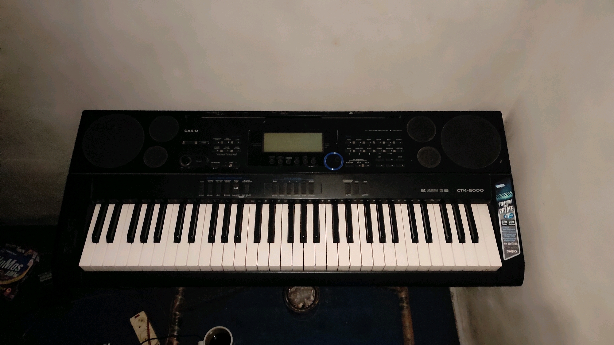 A black Casio CTK-6000 keyboard is positioned centrally in the frame, angled slightly to the right. The keyboard has white and black keys, a digital display screen, and various buttons and knobs. To the left of the keyboard, a small portion of a packet with colorful graphics and text is visible, suggesting it's some form of consumer product. Below the keyboard, dark blue flooring is apparent. In the bottom left corner, a white electrical adapter with wires is partially visible. To the right of the adapter, a white mug with dark liquid, likely coffee, is present. The background is a plain, light-colored wall. The overall scene appears to be an indoor setting, possibly a room.