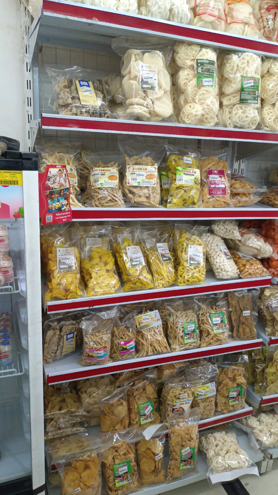 The image shows a retail display of Indonesian snacks in plastic packaging, arranged on white shelves with red trim. The products appear to be various types of crackers and crisps. On the left side of the image, part of a white refrigerator or display case is visible. The shelving unit is filled with numerous clear plastic bags, each containing a different type of snack. Some bags have colorful labels with text in Indonesian.
Based on the context of the products and their packaging, this scene is likely in a small grocery store, minimarket, or a traditional market stall in Indonesia. The specific mention of Padang Panjang, Indonesia, places this scene in a city on the island of Sumatra. The image focuses on the products for sale, with no people clearly visible in a transactional interaction. The environment is clean and organized, suggesting a commercial setting.