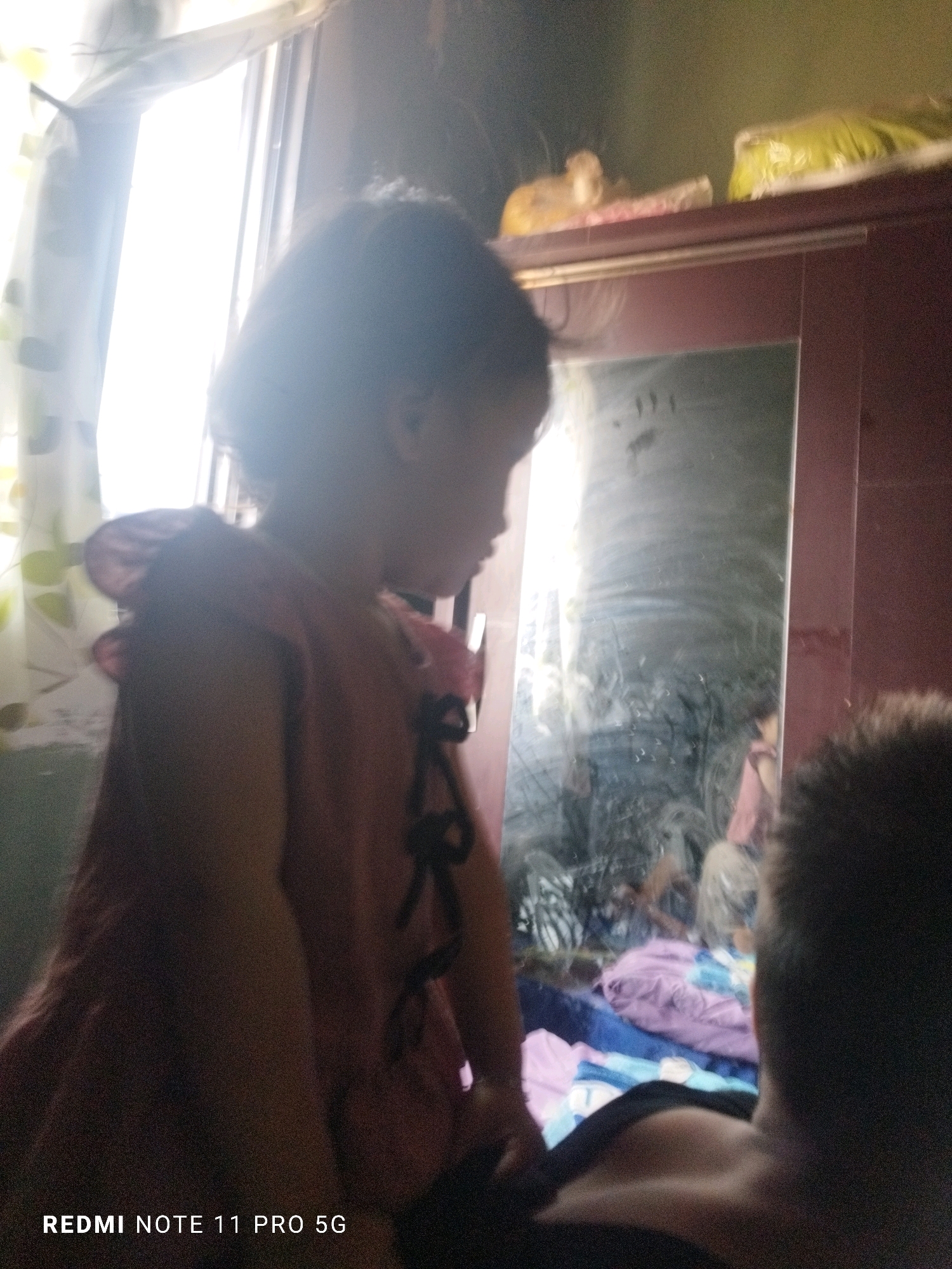 An indoor scene depicts a young child in the foreground, facing right, with a portion of an adult's head visible in the lower right. The child wears a reddish-pink garment with ruffled shoulders and black bows. Behind the child, a tall, dark-colored cabinet with a mirror is present. The mirror reflects a bright window with sheer curtains, the cabinet's top surface, and various items on it, including what appears to be plastic-wrapped objects and a small figurine. The room seems to be a bedroom, with bedding visible in the reflection of the mirror. The lighting suggests it is daytime, with light streaming in from the window. The image is tagged "REDMI NOTE 11 PRO 5G". The location is identified as Kaduhejo, Indonesia.