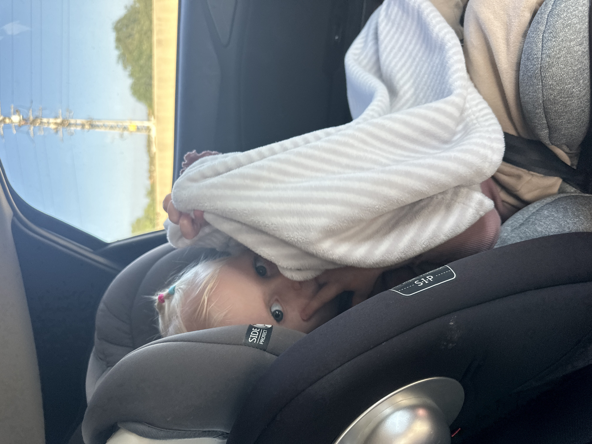 The image, rotated 90 degrees clockwise, depicts a young child seated in a grey and black car seat inside a vehicle. The child, with blonde hair styled with small pink and blue hair ties, has wide blue eyes and is looking directly at the camera. They are partially covered by a soft, light-colored blanket with white and light grey stripes, peeking out from underneath it in a playful manner.
The car seat features visible text, including "SIDE PROTECT" on the headrest and "S.I.P" on a label. A metallic cup or container is partially visible in the lower right corner of the frame. Through the car window, a bright, clear sky indicates daytime, with blurred green foliage (suggesting trees) and what appears to be a power pole or utility lines in the distance. The setting suggests the car is either stationary or in motion. The child's expression conveys curiosity and playfulness. The location context for this scene is Rishpon, Israel.