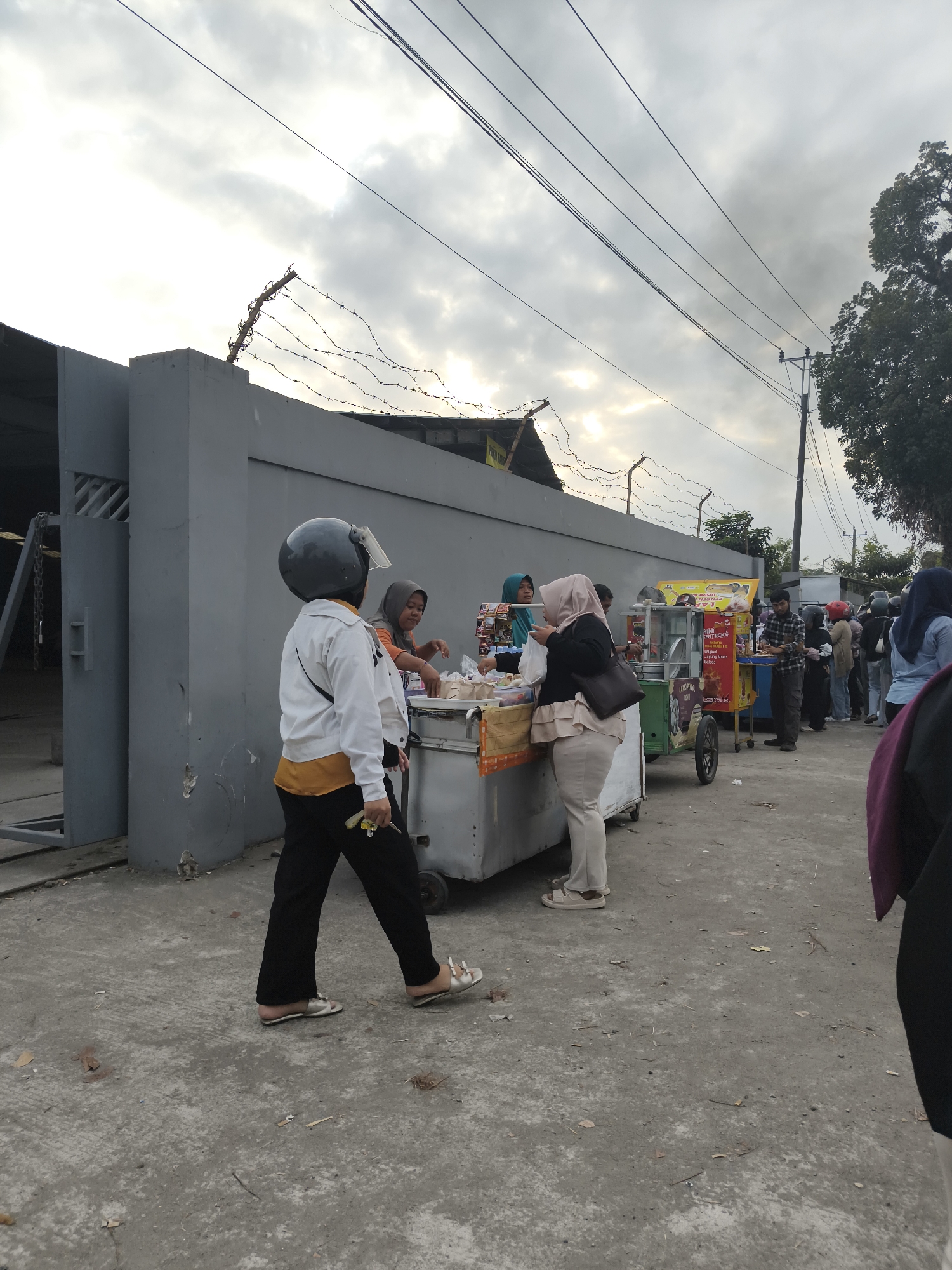 In Bukateja, Indonesia, a street scene unfolds with a line of people waiting to purchase food from vendors. In the foreground, a person wearing a black helmet, white jacket, and black pants walks away from the camera. To their left, a grey concrete wall with a metal gate is visible. Behind the wall, barbed wire is strung across its top. Further down the street, two food carts are set up, with vendors serving customers. Several people, mostly women wearing hijabs, are clustered around the carts. In the background, a utility pole with power lines stretches across the cloudy sky. The ground is paved with concrete.