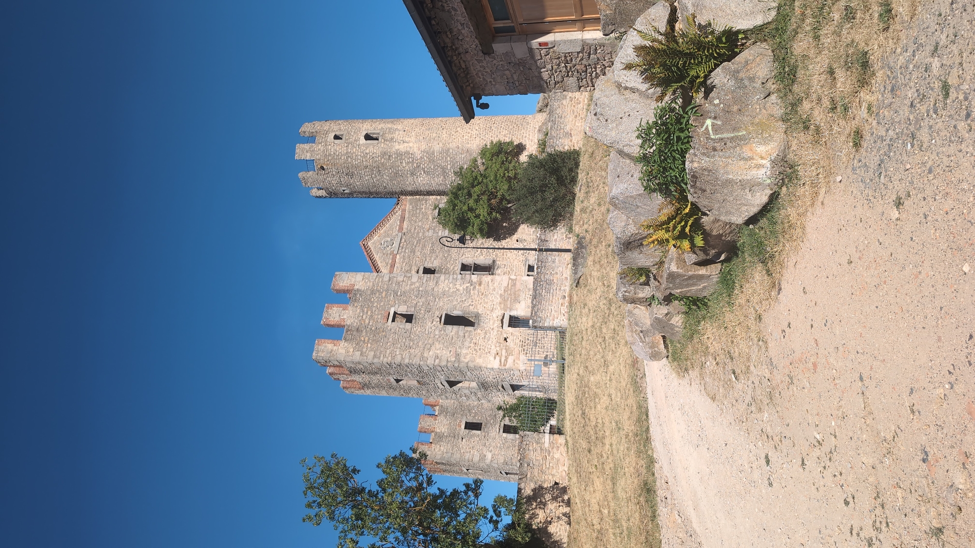 A daytime, low-angle, exterior, full shot shows a medieval castle constructed of stone and brick under a clear blue sky. The castle is situated on a grassy hill, with a dirt path leading up to it. In the foreground, several large rocks are piled together, with some greenery growing between them. To the right of the castle, a section of a stone building with a wooden door is visible. The castle itself features multiple towers and battlements, with numerous empty window openings. A tree with green foliage stands to the left of the castle, and another smaller, bush-like tree is situated in front of the castle walls. A lamppost with a curved arm stands near the center of the image, adjacent to the castle. There is no visible evidence of human activity or presence. This is a view of the Château de Chambles, located in Chambles, France.