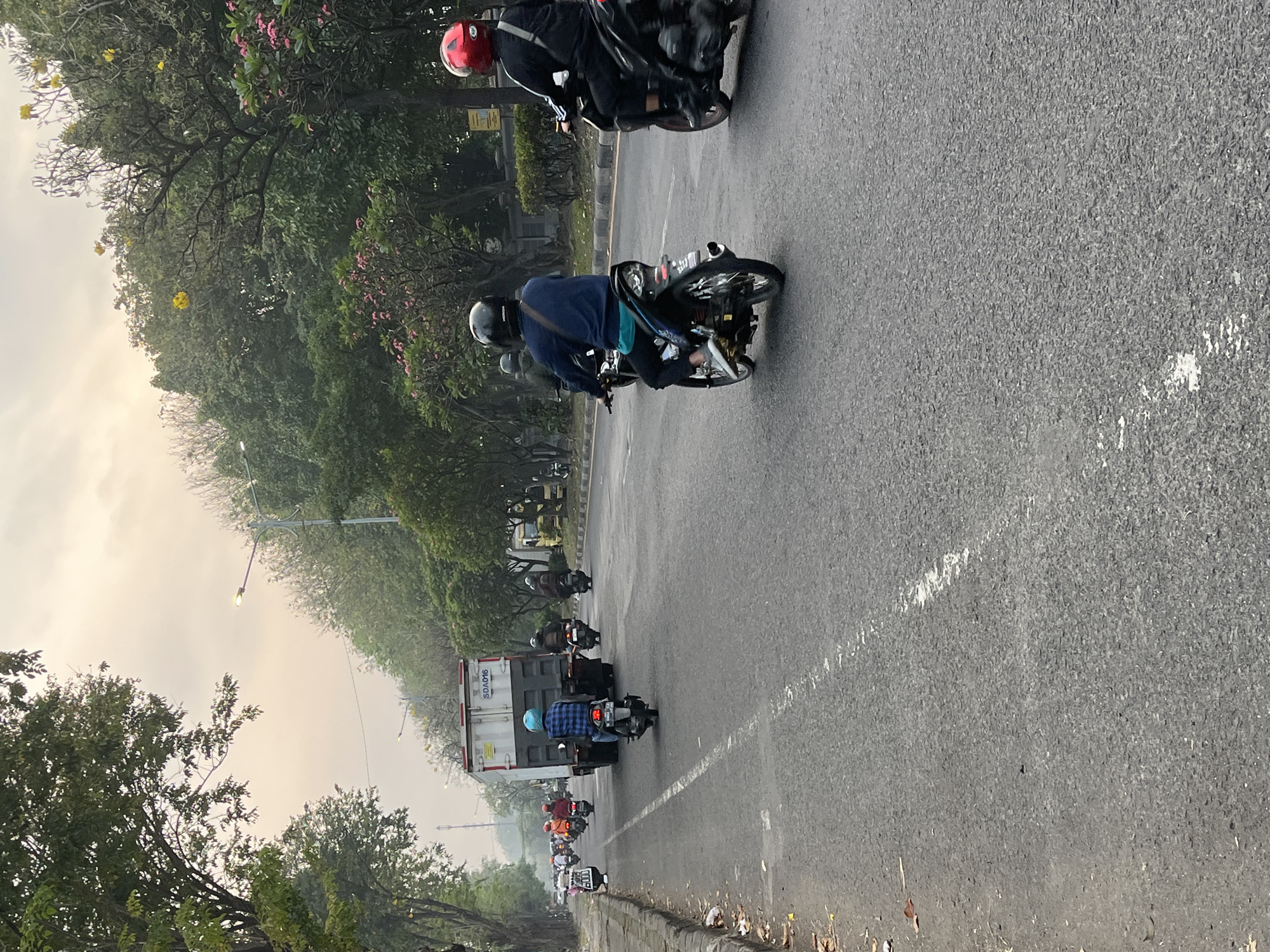 The image depicts a road scene in Sumorame, Indonesia, characterized by a cloudy sky and lush green trees lining the street. Multiple individuals are riding motorcycles, some with passengers. In the foreground, a motorcycle rider wearing a blue checkered shirt and a light blue helmet is seen from behind, moving away from the camera. To the right, another motorcyclist with a red helmet is also visible. Further down the road, a large truck with "SDA016" visible on its side is in motion. The road surface is asphalt with visible white lane markings. The overall atmosphere suggests a typical day in a moderately busy urban or suburban area.
